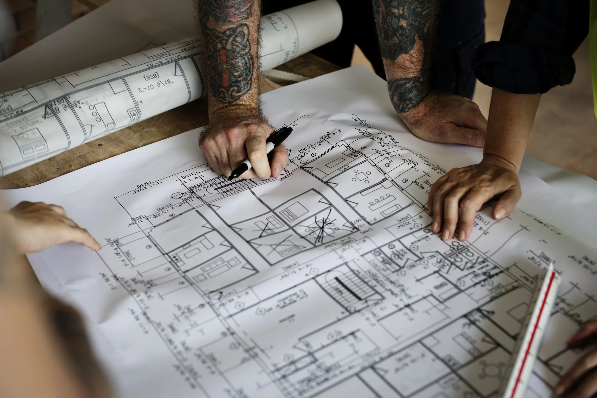 Architects or engineers reviewing detailed blueprints or floor plans on a large table with rolled-up architectural drawings in the background.
