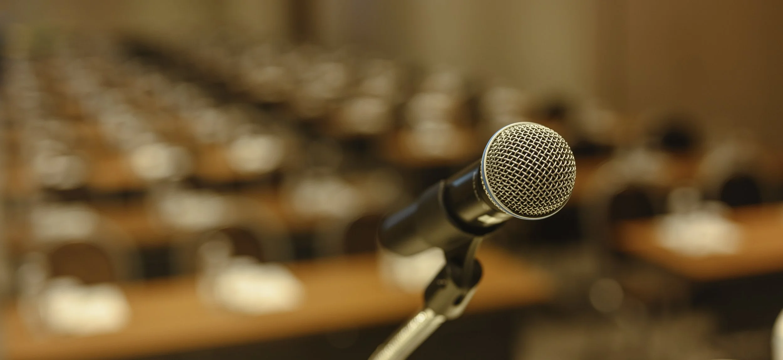 A close-up of a microphone on a stand, with a blurred background of sound equipment or audio mixing controls.