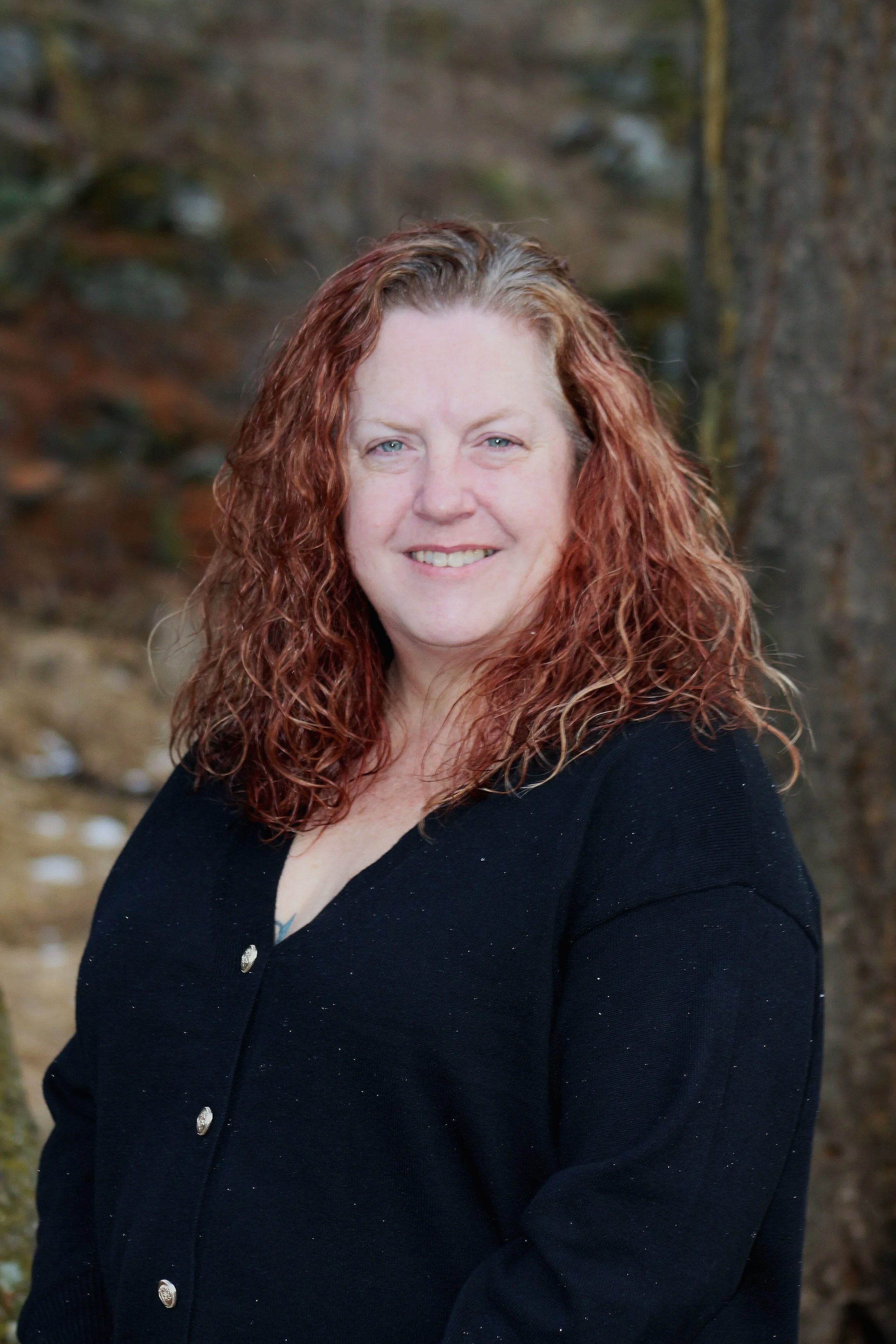 A woman with curly red hair and blue eyes smiling, wearing a black cardigan with silver buttons, standing outdoors in front of blurred trees and water.