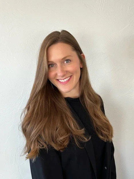 A woman with long brown hair, smiling, wearing a black top, standing against a plain white wall.