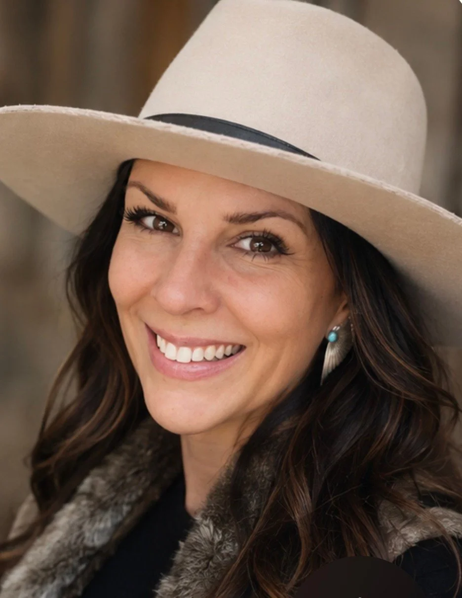 A woman smiling, wearing a wide beige hat with a black band, a black top, and a coat with a fur collar. She has long wavy brown hair and blue earrings.