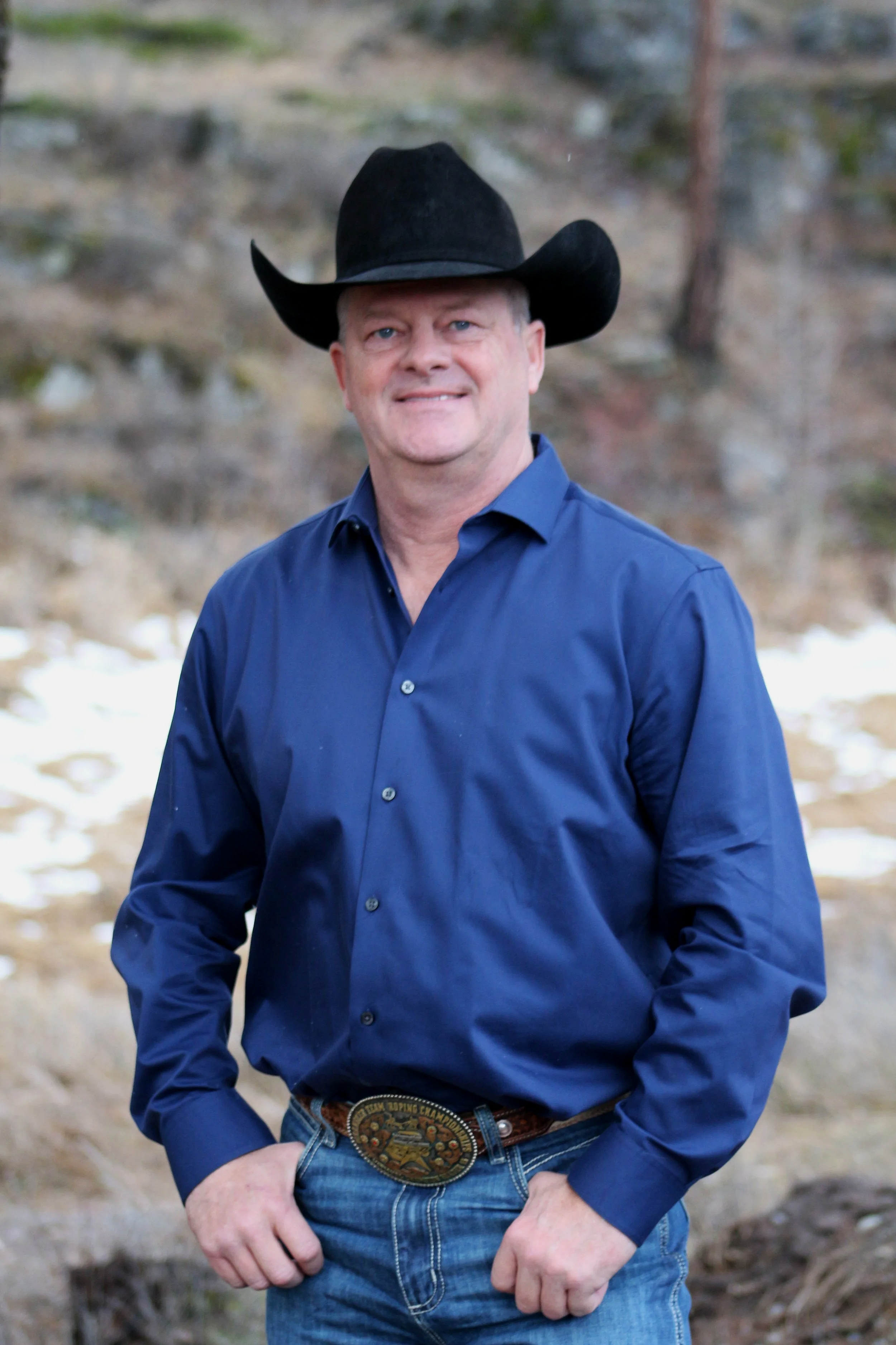 A man wearing a black cowboy hat, blue dress shirt, jeans, and a Western-style belt buckle stands outdoors in a natural setting with trees and snow on the ground, smiling at the camera.
