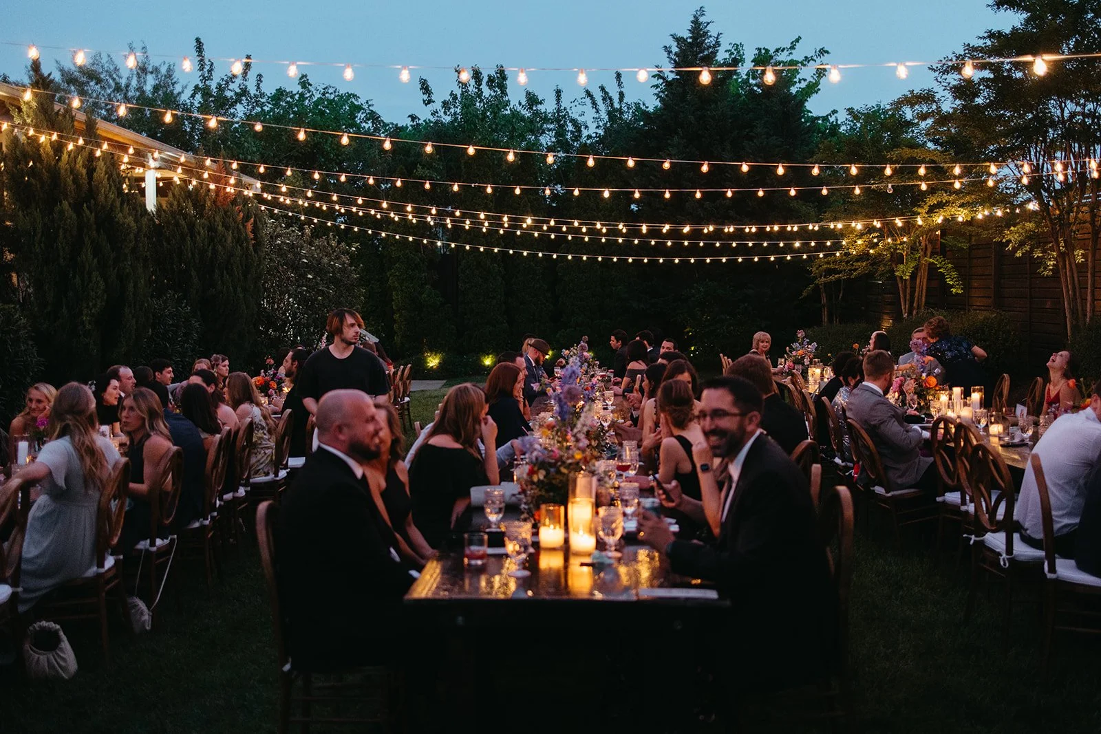 An Gorgeous outdoor evening dinner party in the Garden at the Cordelle in Downtown Nashville with long table set with candles, flowers, and glassware, illuminated by string lights and surrounded by trees and people dressed formally.