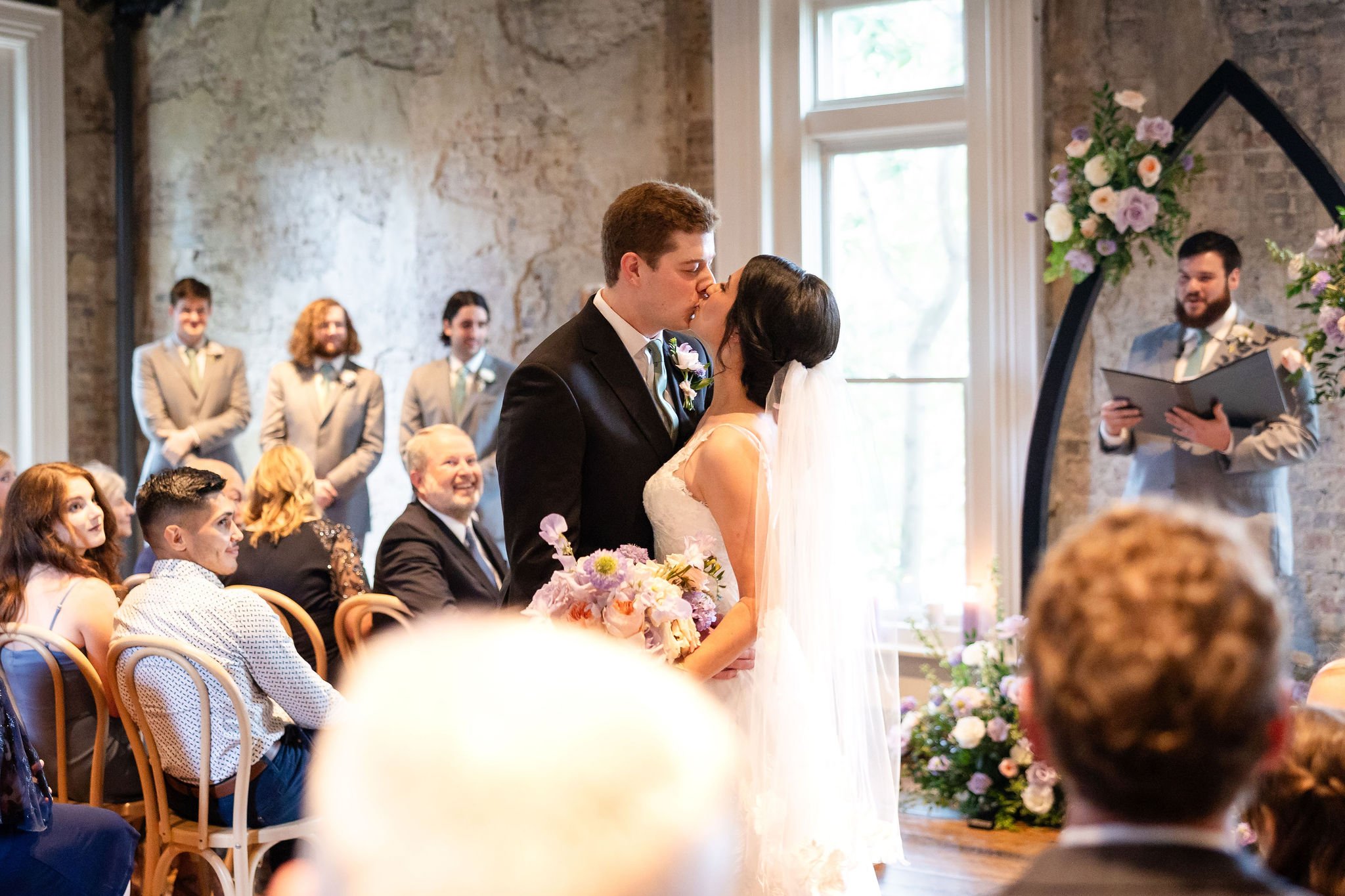 Wedding ceremony in the Victorian at the Cordelle with bride and groom kissing inside a rustic brick venue with large windows, floral arrangements, and seated guests watching.