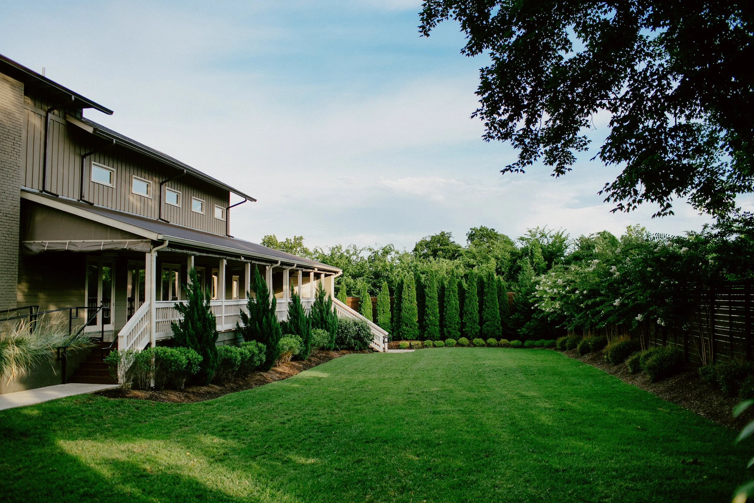 The Cordelle lawn with a well-maintained green lawn, various bushes, and tall hedges. There is a house with a porch, stairs, and multiple windows on the upper level. Trees and a wooden fence surround the area, and the sky is partly cloudy.