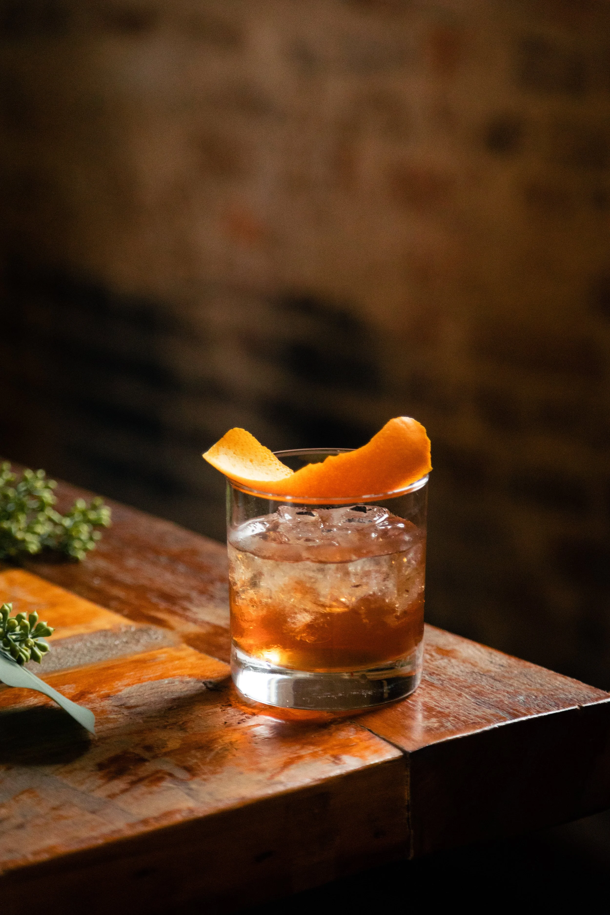 Cordelle Bar Experience Event Bar Nashville A glass of an amber-colored cocktail with ice and an orange peel garnish on top, placed on a wooden bar table.