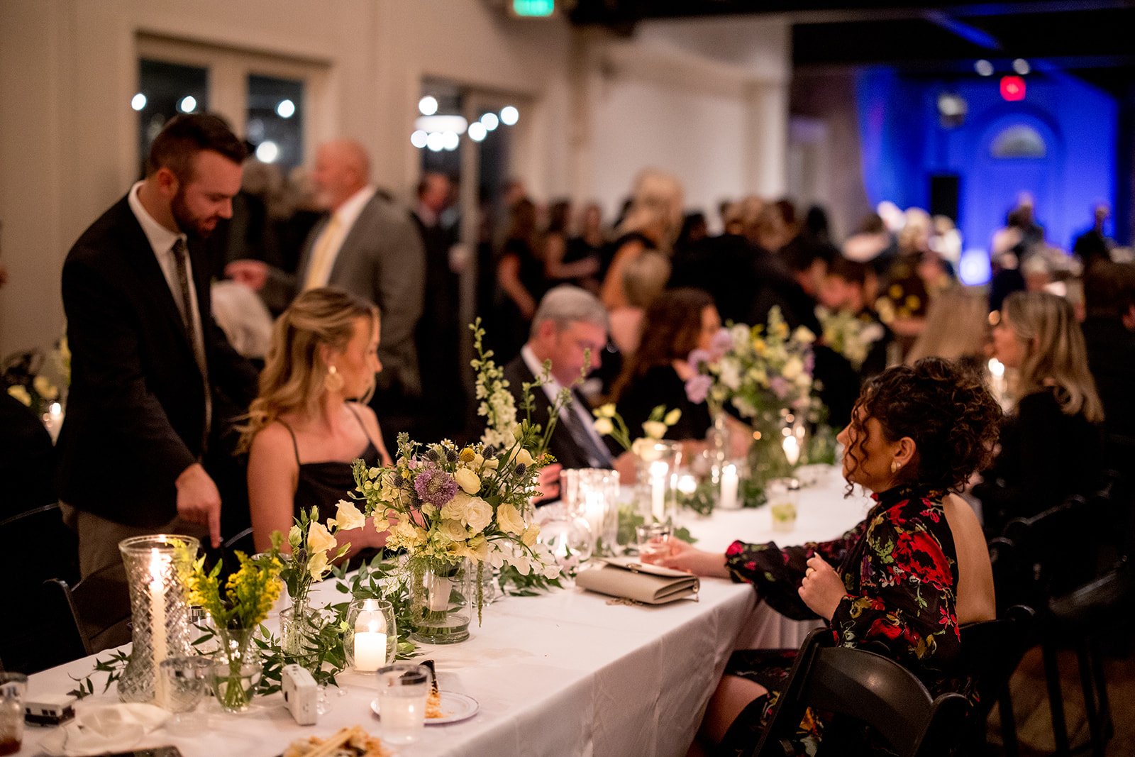 People seated at a long banquet table decorated with flowers and candles at an elegant event or dinner for a reception for the Cordelle.