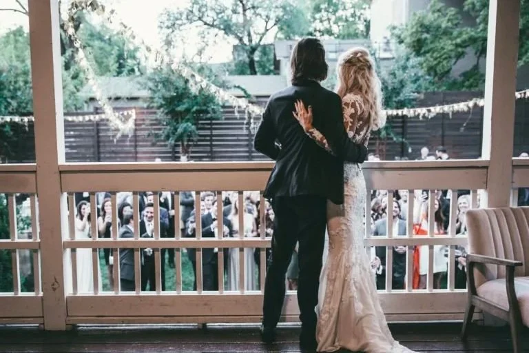 A couple, dressed in wedding attire, stands on a porch overlooking a crowd at an outdoor wedding reception greeting guests in the Gorgeous garden at the Cordelle.