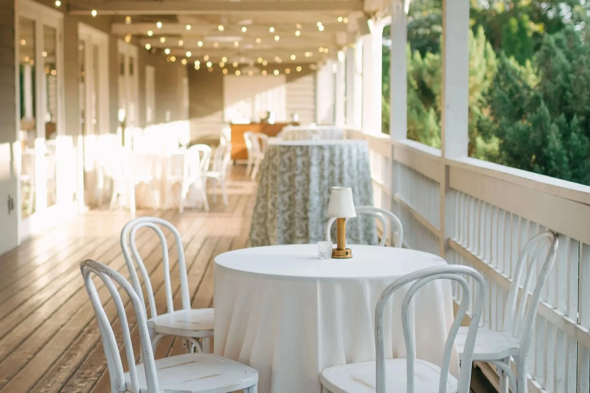 Cozy covered porch with white round table, white chairs, a small lamp, and hanging string lights, overlooking green trees outside in downtown Nashville.