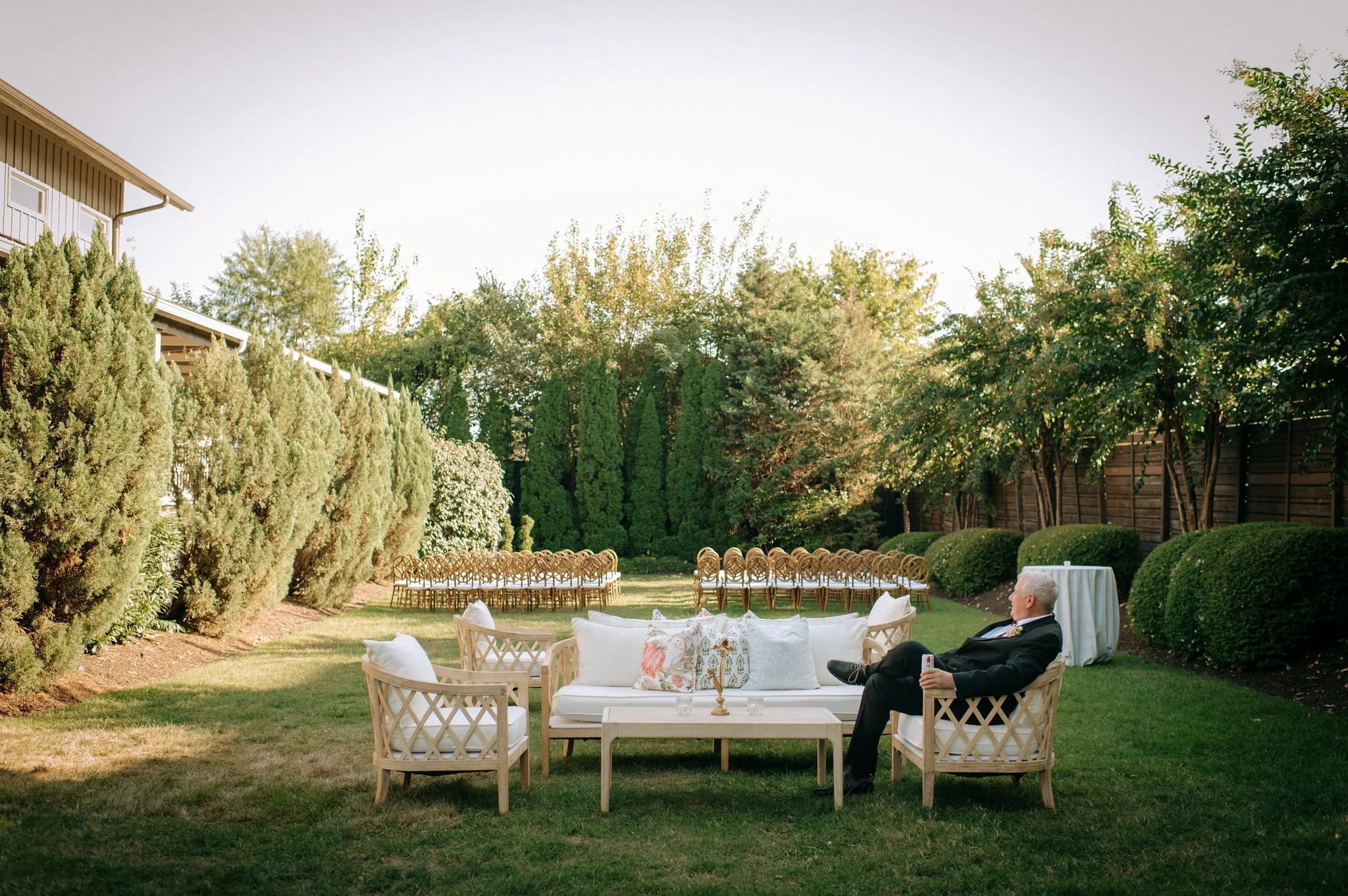 Outdoor garden setting with white furniture and a man in a tuxedo sitting on a chair, surrounded by trees and bushes, with chairs arranged for an event and a table with a white tablecloth.