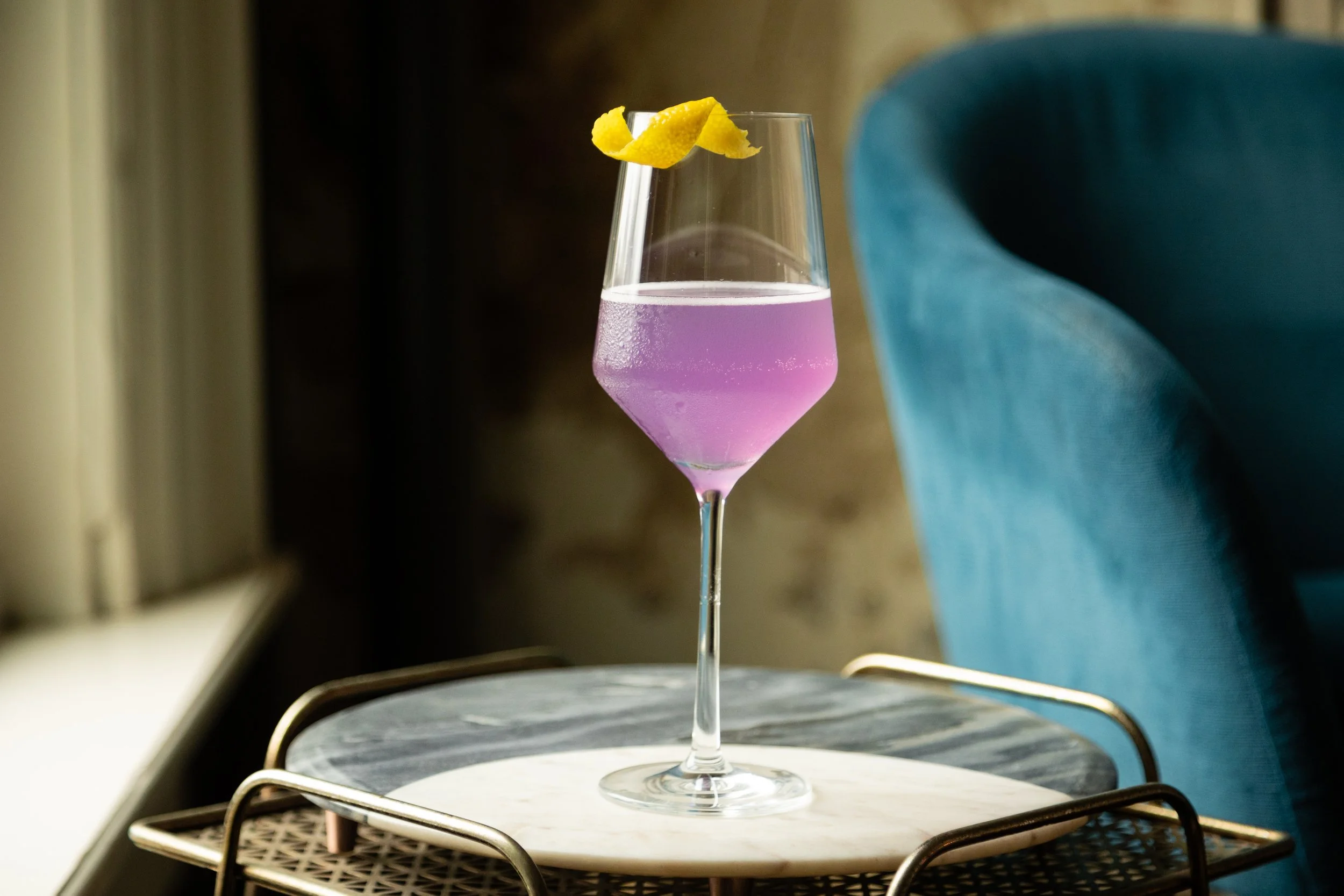 Cordelle Signature cocktail in a tall, slender glass with pink and clear layers, garnished with a lemon twist, placed on a marble serving tray with a tray underneath, beside a teal chair.