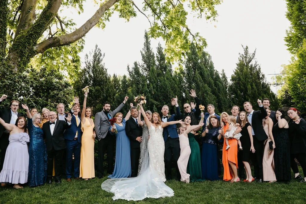 A group of people celebrating a wedding outdoors. The bride and groom are in the center, with the bride in a white wedding dress and the groom in a dark suit. The guests are dressed in colorful formal attire, standing on a lawn with tall trees and gr