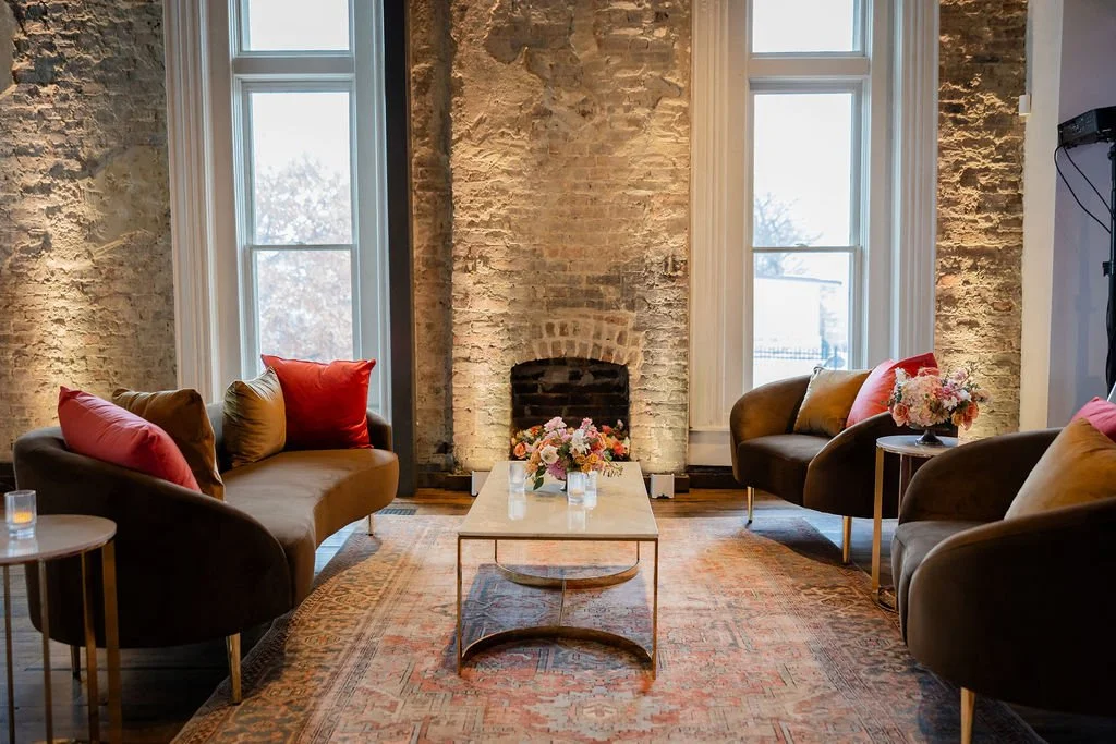 The Victorian at the Cordelle with exposed brick walls, two tall windows, and a brick fireplace. There are four brown velvet armchairs with colorful pillows around a rectangular coffee table decorated with a floral arrangement.