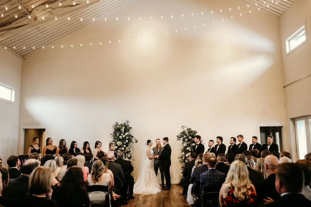Indoor wedding ceremony at award winning venue, the Cordelle, with bride and groom holding hands, surrounded by bridesmaids and groomsmen, guests seated, and white string lights hanging from ceiling.