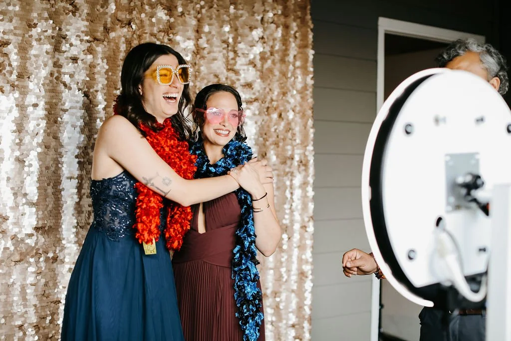 Wedding reception a the Cordelle photo booth Two women standing together, smiling for a photo, with party accessories like leis and glasses, in front of a shiny gold backdrop, while a person takes their picture with a camera.
