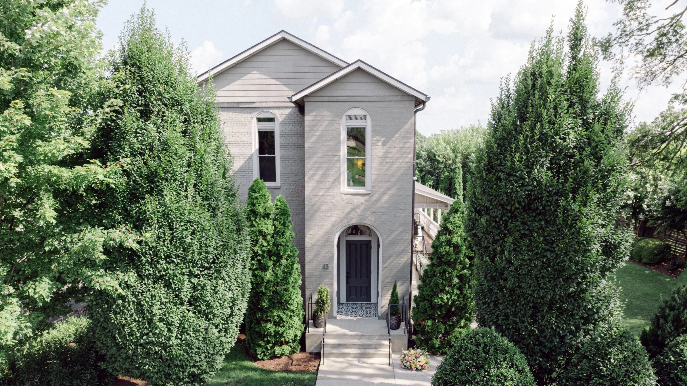 Exterior of the Cordelle in Downtown Nashville wedding venue and corporate event space A two-story house with gray brick exterior and pointed roof, partially obscured by tall green trees with a lush yard, front steps, and a black door.