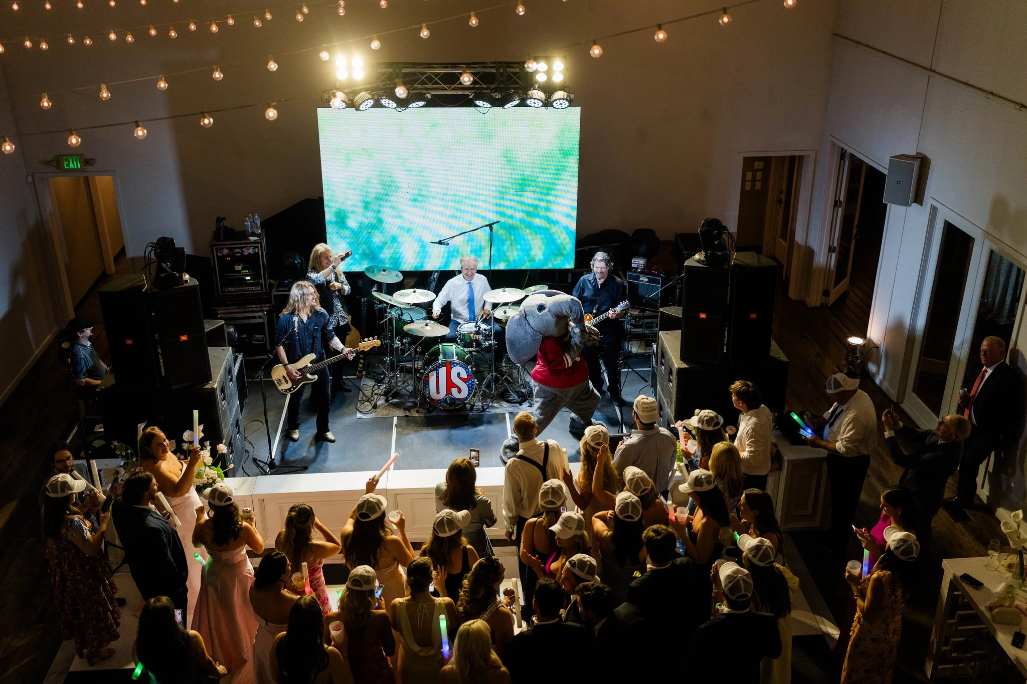 A band performing live on stage in the Great Hall at the Cordelle for a Corporate event with a large digital screen behind them, surrounded by an audience in a decorated indoor venue with string lights overhead.