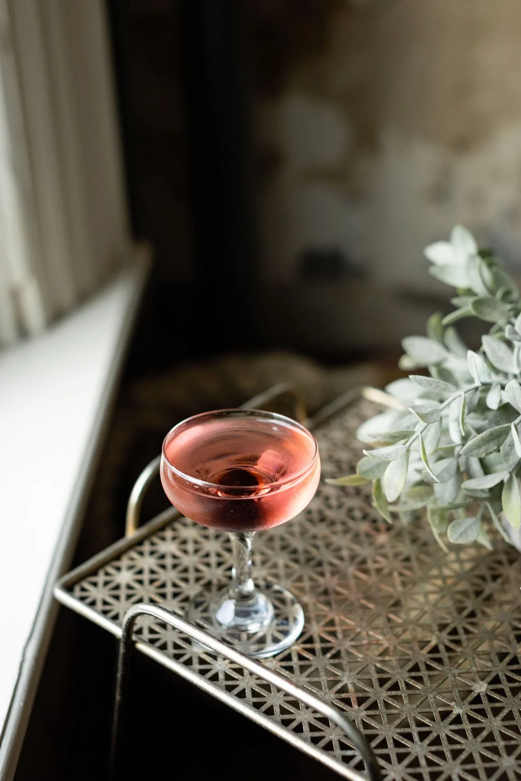 A Signature cocktail in the Suite at the Cordelle with glass of pink cocktail on a decorative metal tray next to a green plant, near a window.