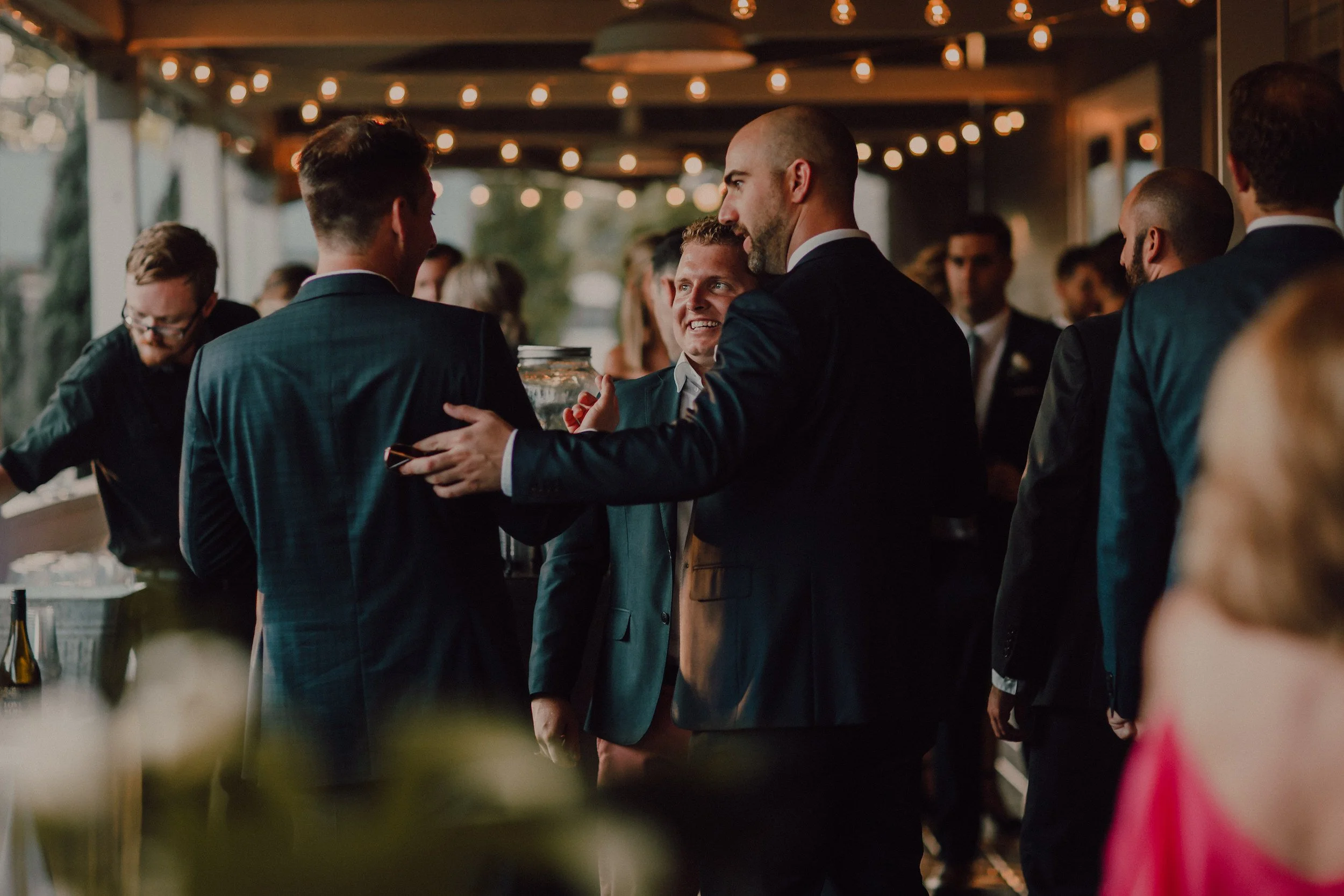 Corporate event gathering a group of men in suits at a social event, chatting and smiling in a warmly lit venue with string lights overhead.