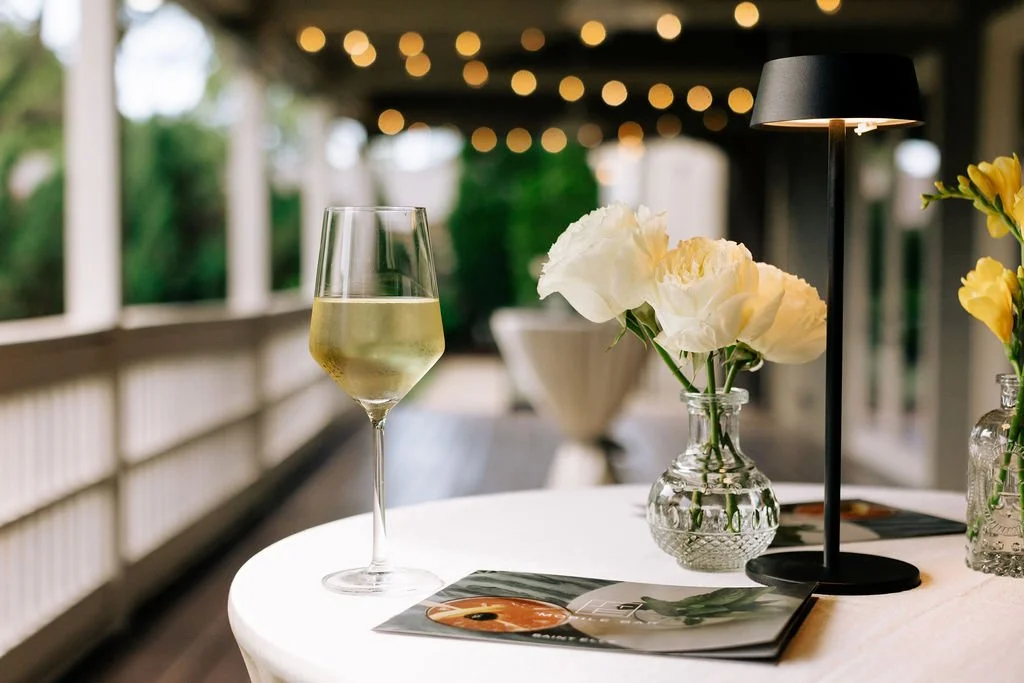 A glass of white wine on the Covered Porch at the Cordelle in downtown Nashville, a small table lamp, and vases with white and yellow flowers on a table