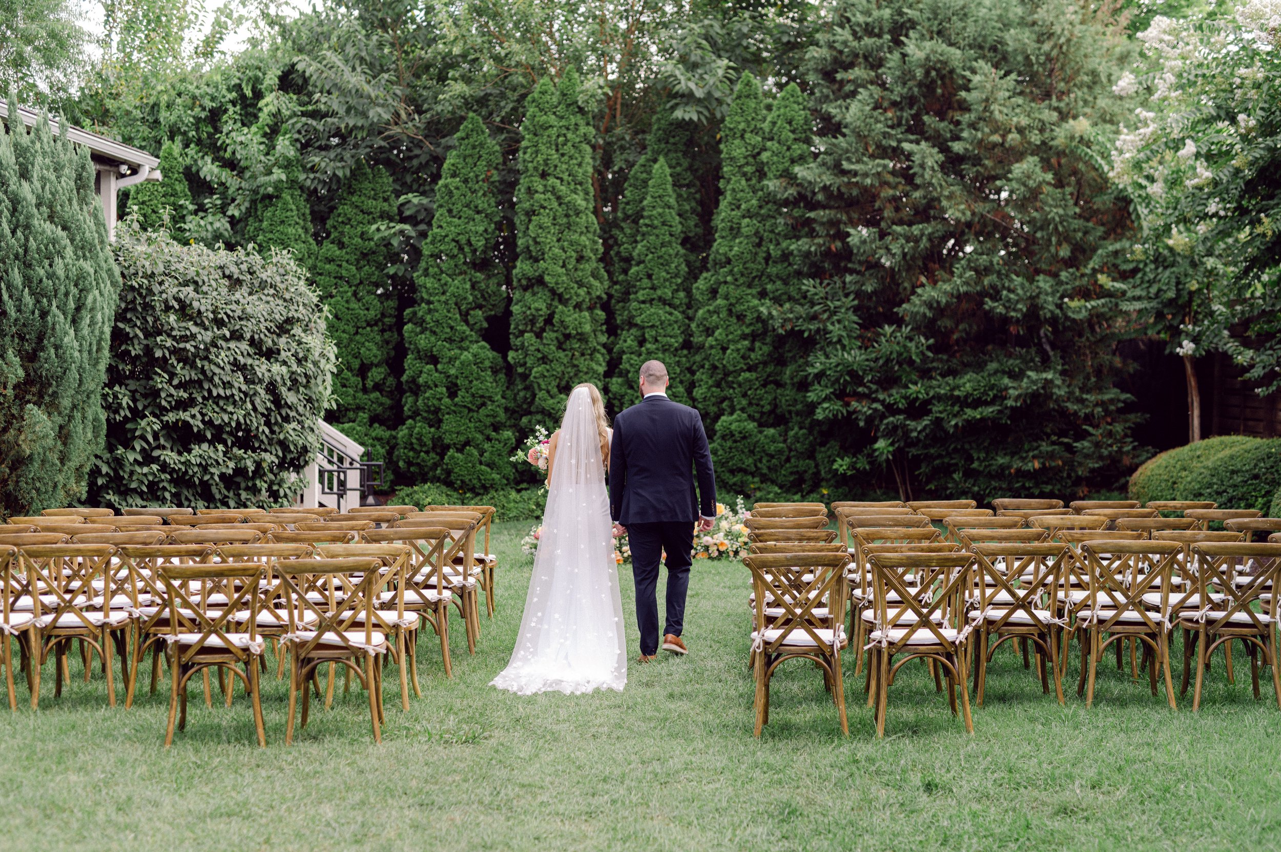 A bride and groom walking down the aisle at an outdoor wedding ceremony in a garden in downtown Nashville surrounded by rows of chairs and lush green trees.