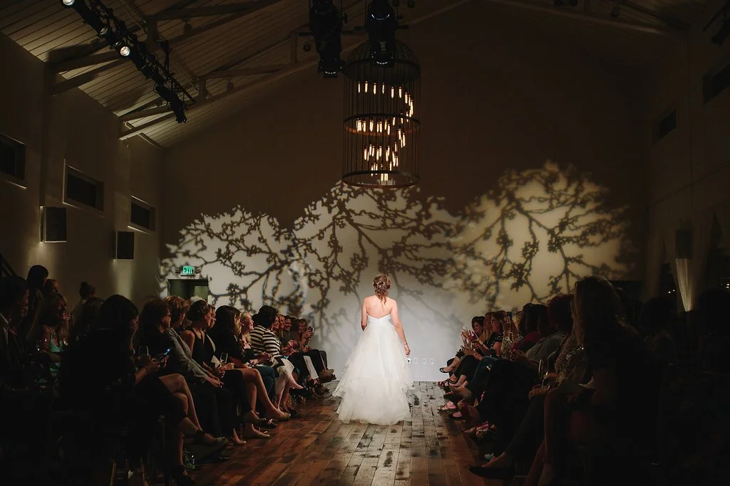 Fashion show in Nashville at the Cordelle A woman in a white wedding dress walking down a runway with an audience on both sides, shadow of tree branches cast on the wall behind her