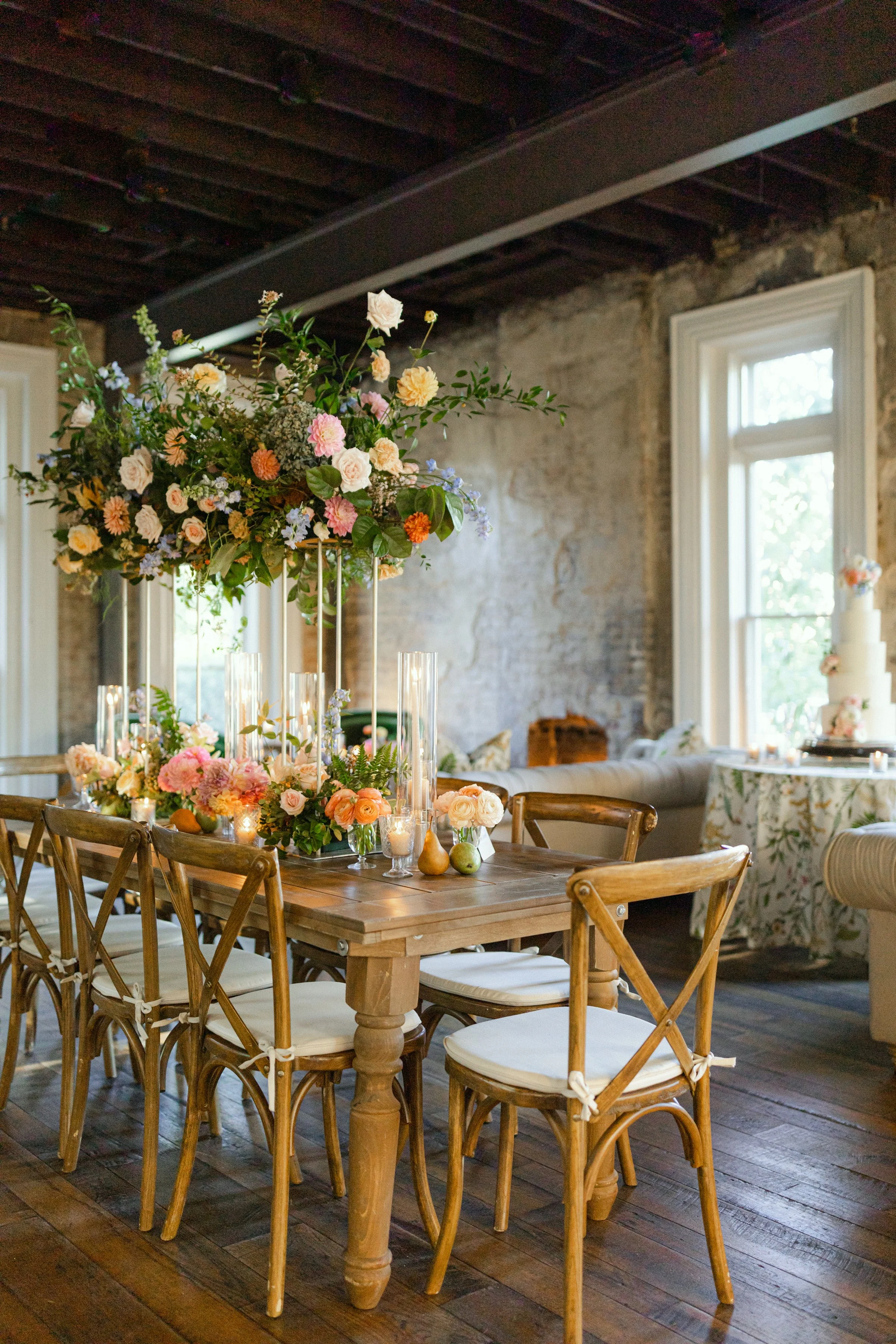 Reception set-up at the Cordelle with A wooden dining table decorated with a large colorful floral centerpiece, surrounded by wooden chairs with white cushions, in a rustic-style room with exposed brick walls and large windows.