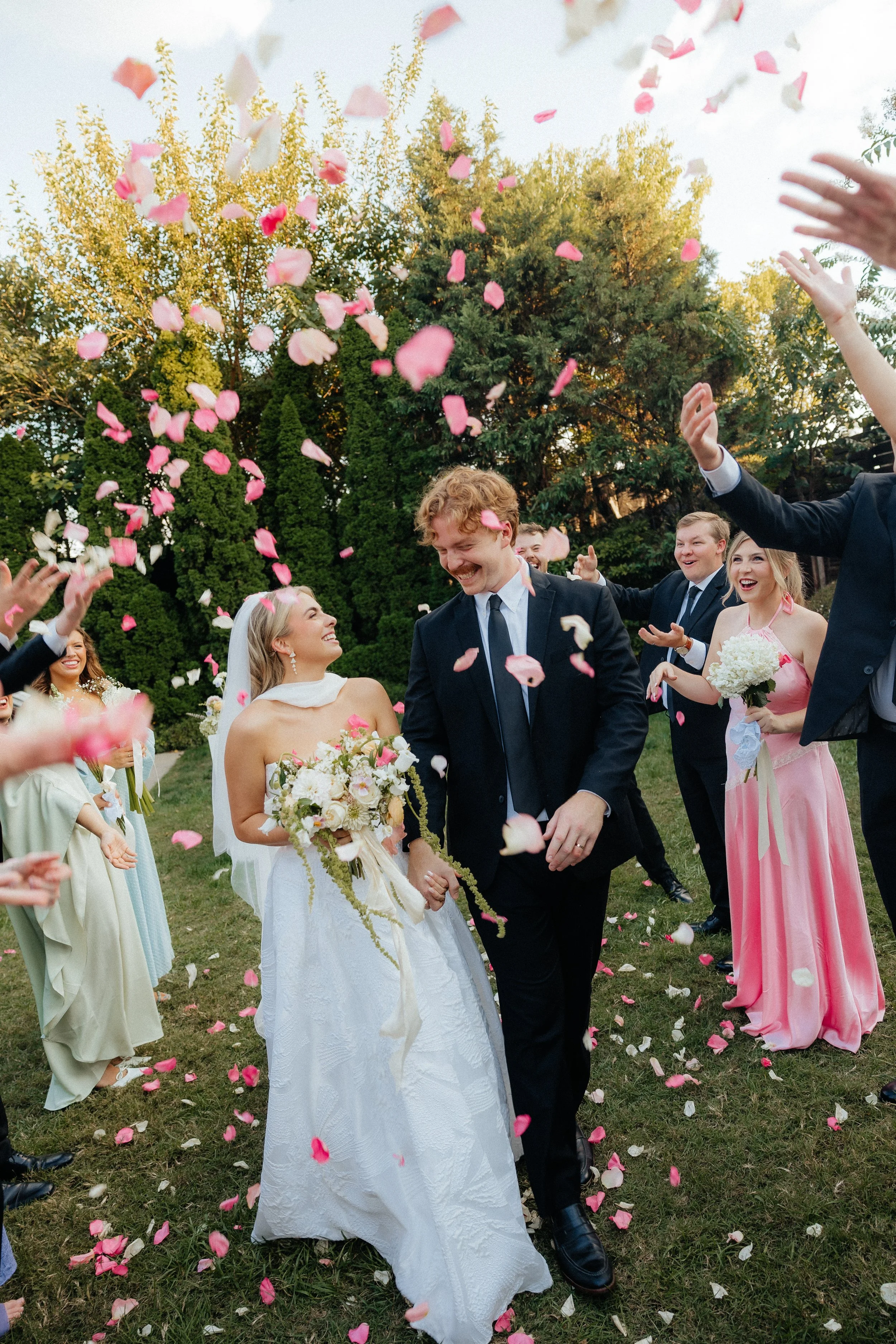 A wedding celebration in the Garden a the Cordelle with a bride and groom walking hand in hand while confetti is thrown in the air by guests, with lush green trees in the background.
