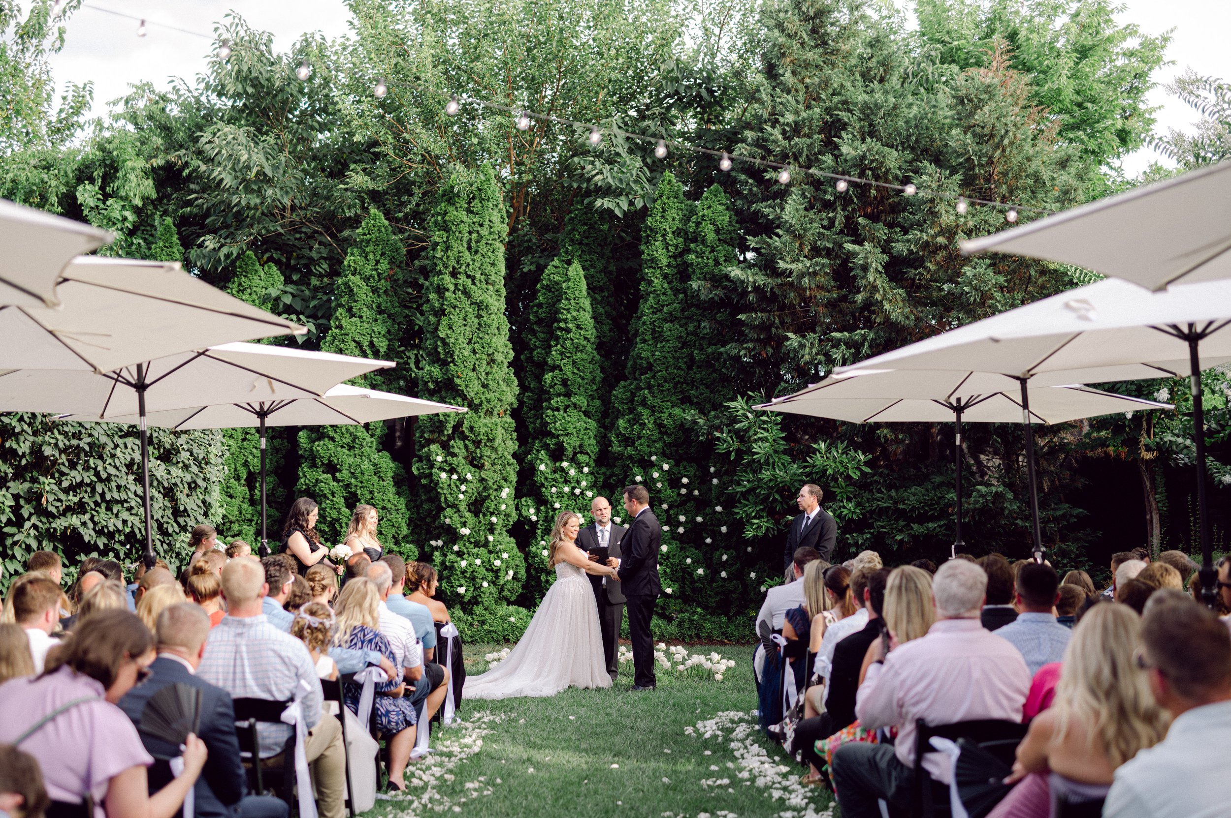 Outdoor Cordelle wedding ceremony with a couple standing before an officiant, surrounded by seated guests, trees, and patio umbrellas.