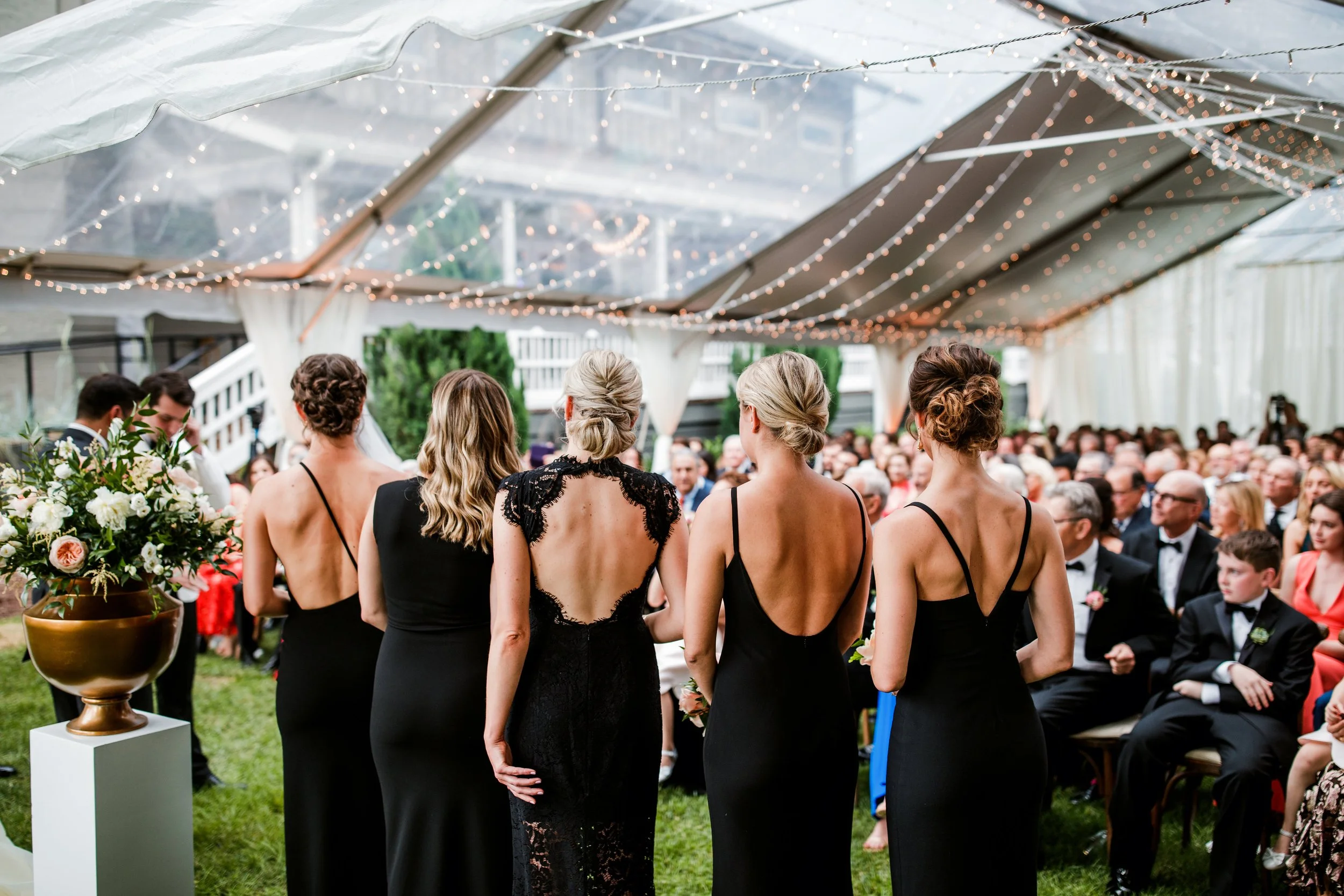 The Cordelle Garden wedding ceremony under a clear tent decorated with string lights, five women with styled blonde hair and wearing elegant black dresses stand with their backs to the camera, facing a seated crowd of wedding guests.