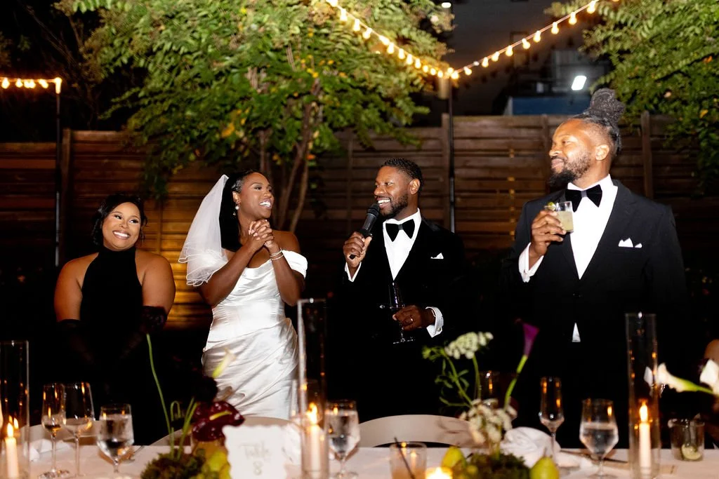A wedding celebration with five people toasting and smiling, outdoors at night, decorated with string lights, flowers, and candles, with a wooden fence and trees in the background.