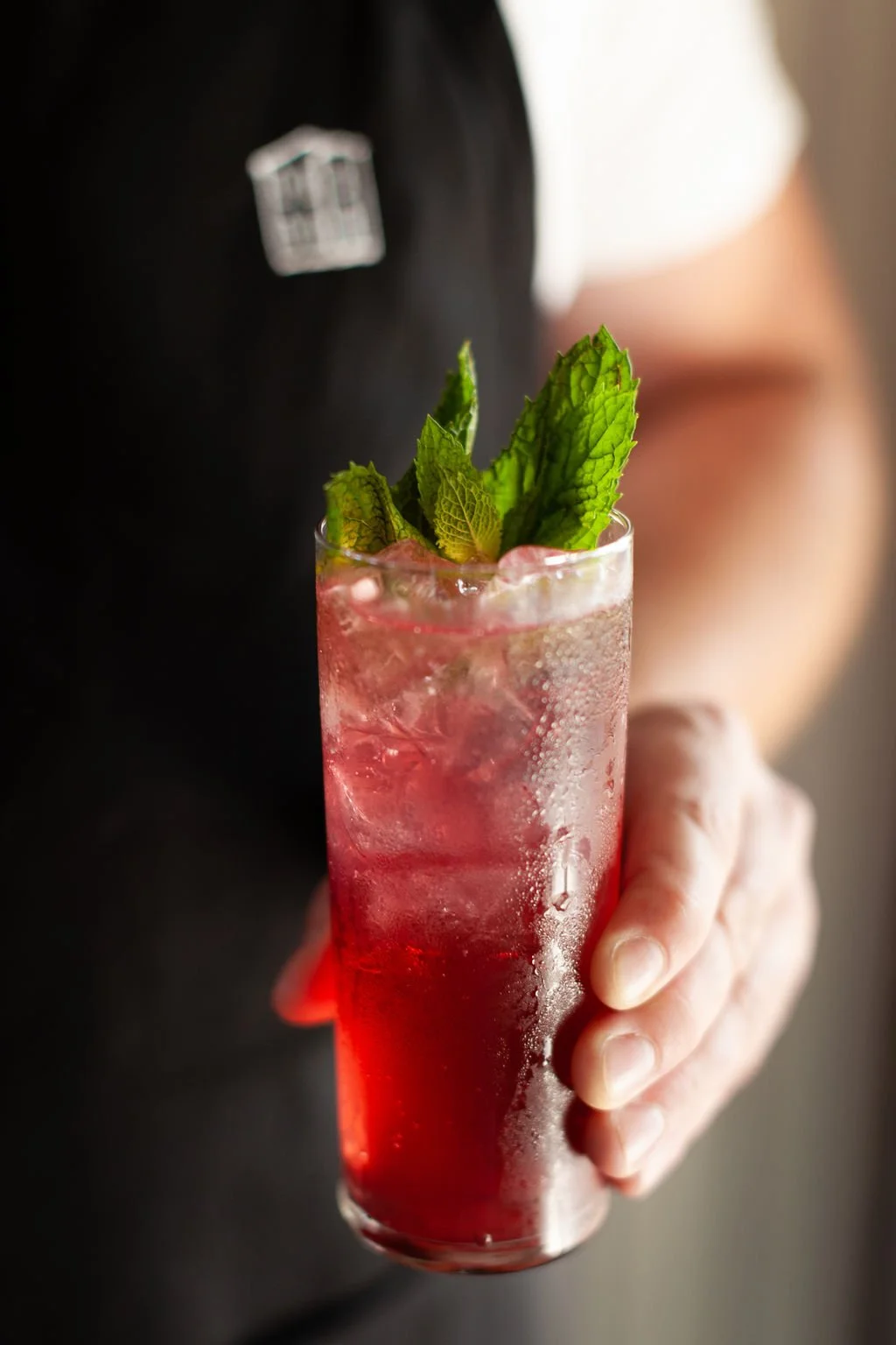 Cordelle Signature Cocktail being served by our beverage team A person holding a tall glass of red cocktail garnished with fresh mint leaves, with condensation on the glass.