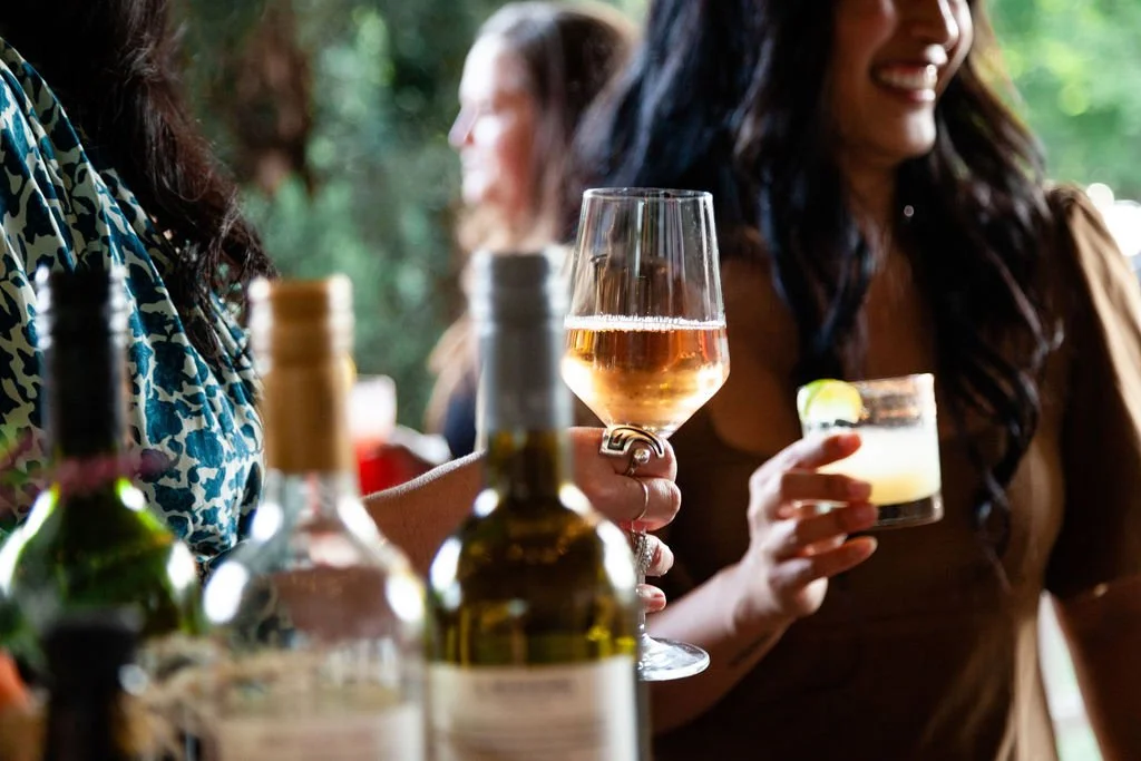 Cocktail hour on the Covered Porch at the Cordelle Women socializing outdoors with drinks, bottles on the table, one woman holding a glass of rosé wine, another holding a cocktail with lime.