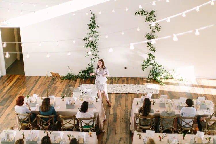 Corporate workshop and product launch at the Cordelle in Nashville A woman giving a presentation to a seated audience in a decorated indoor venue with string lights and greenery.