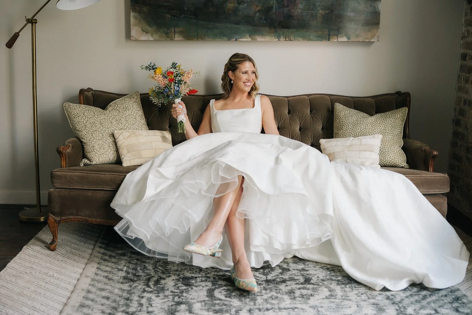 A bride sitting on a vintage brown sofa in the bridal suite holding a bouquet of flowers, smiling in a well-lit room with a large abstract painting above her, patterned throw pillows, a lamp, and a textured area rug.
