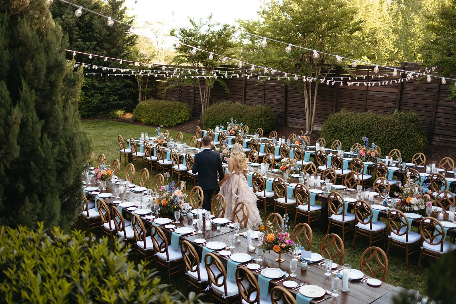 An outdoor wedding reception setup on a lawn with long tables decorated with floral arrangements, surrounded by wooden chairs, under string lights with trees and a wooden fence in the background. A bride and groom walk hand in hand.