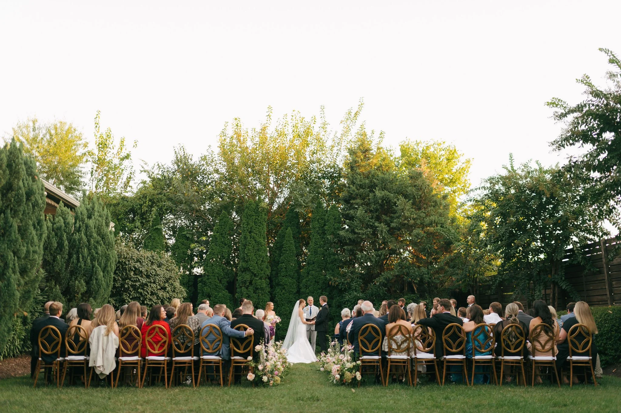 Outdoor wedding ceremony at the Cordelle in Downtown Nashville with seated guests, a bride and groom, officiant, in a garden with trees and a wooden fence.