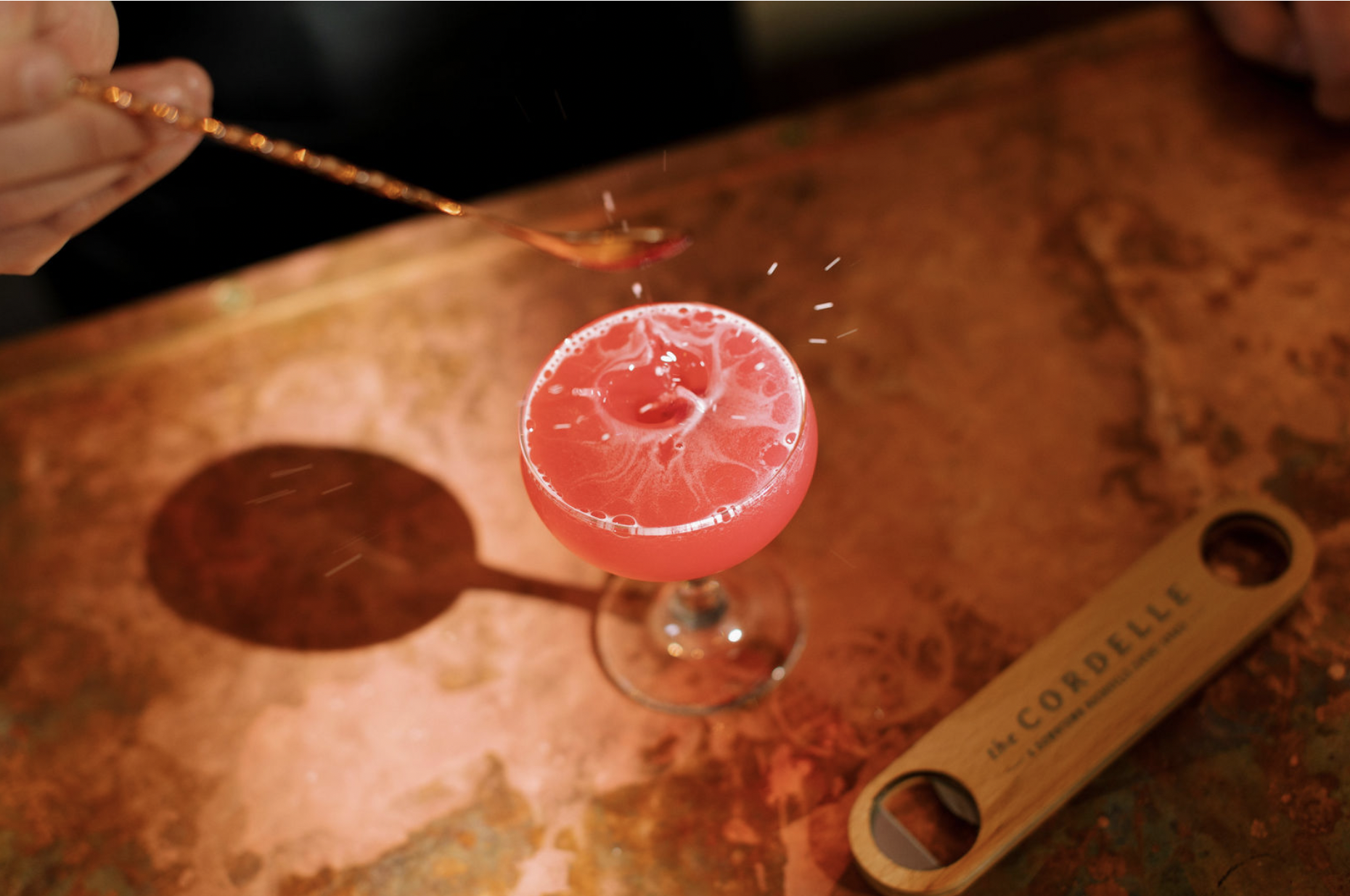Cordelle Bar Experience Event Bar Nashville A pink cocktail in a stemmed glass with a lemon wedge garnish, placed on a textured orange-red countertop, with a hand pouring from a copper spoon into the drink and a wooden tasting spoon nearby.