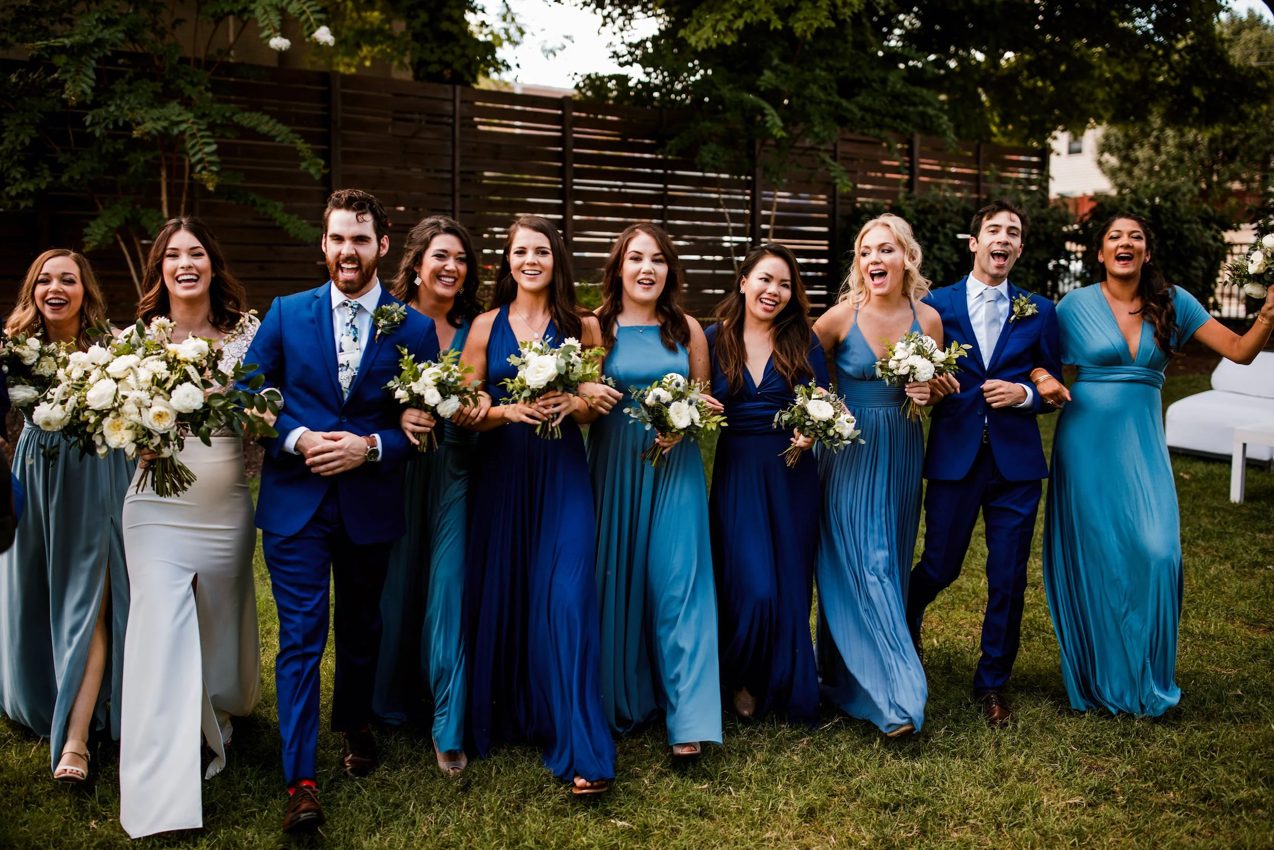 A Cordelle wedding party in formal attire, mostly women in blue dresses and men in suits, walking and smiling outdoors at a wedding or celebration in the Garden in downtown Nashville.