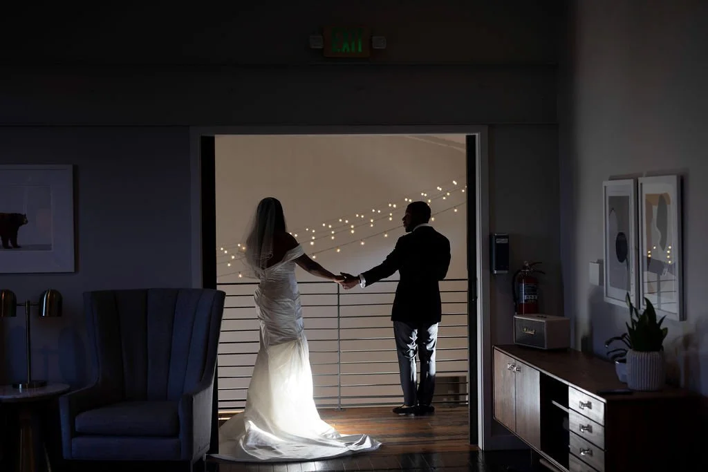 Silhouette of a bride and groom holding hands on the loft balcony with string lights in the background, looking over the Great Hall at the Cordelle.