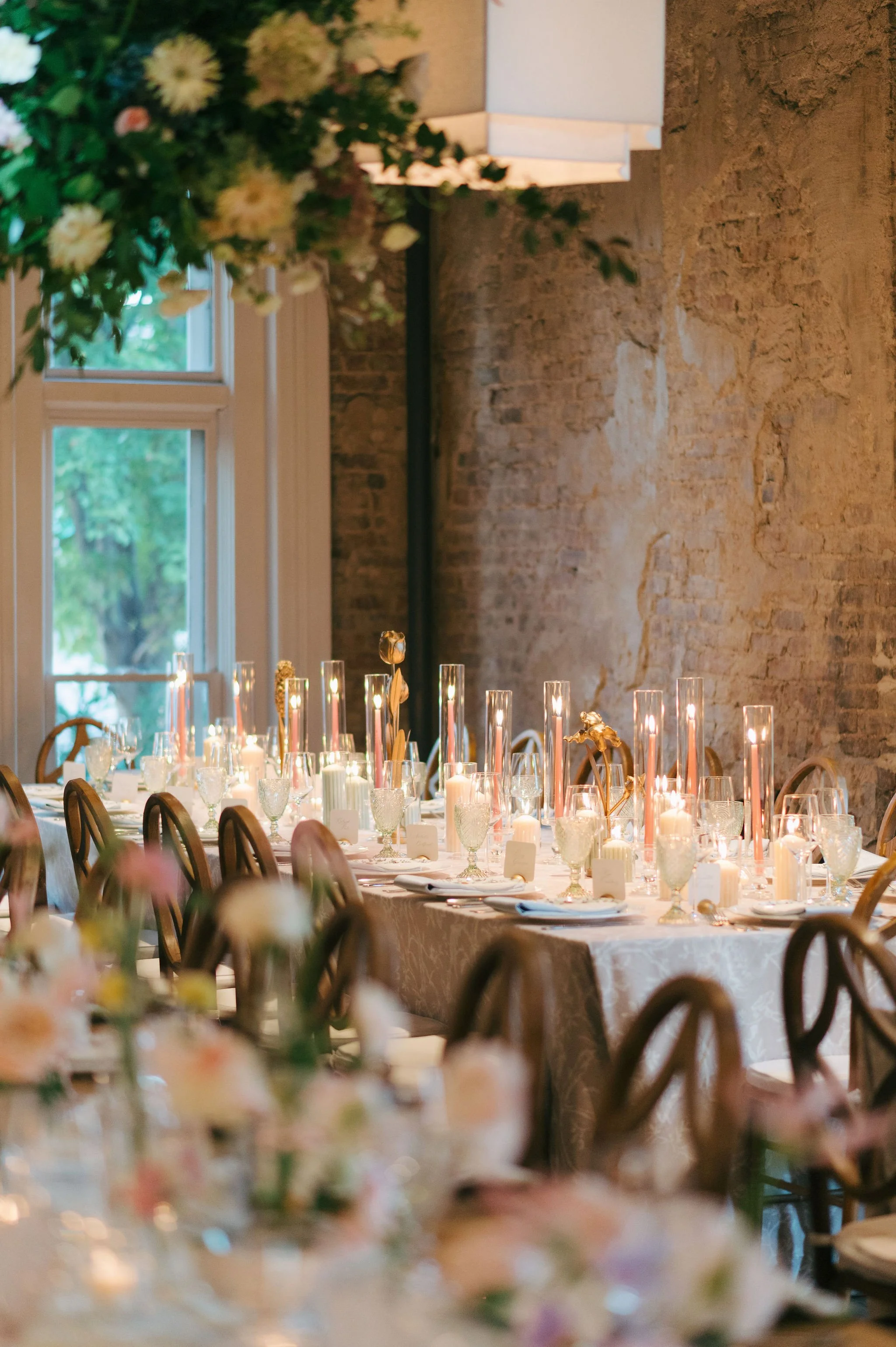 A beautifully decorated dining table set for a formal event or celebration with candles, floral arrangements, and elegant tableware in a rustic room with a brick wall and large windows.