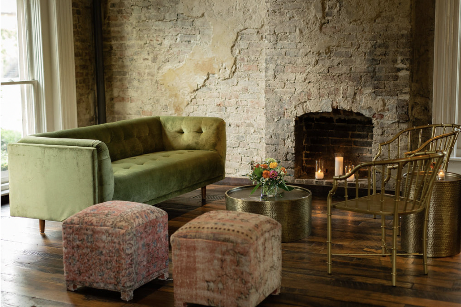 the Victorian at the Cordelle with exposed brick wall, green velvet sofa, two patterned ottomans, gold chairs, a round gold coffee table with a flower arrangement, and a fireplace with lit candles.