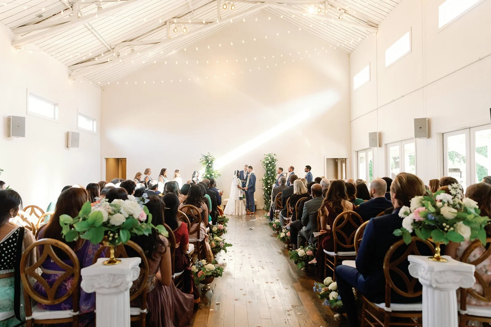 Indoor wedding ceremony in the Great Hall at the Cordelle with bride and groom exchanging vows on a wooden aisle flanked by floral arrangements and floral pillars. 