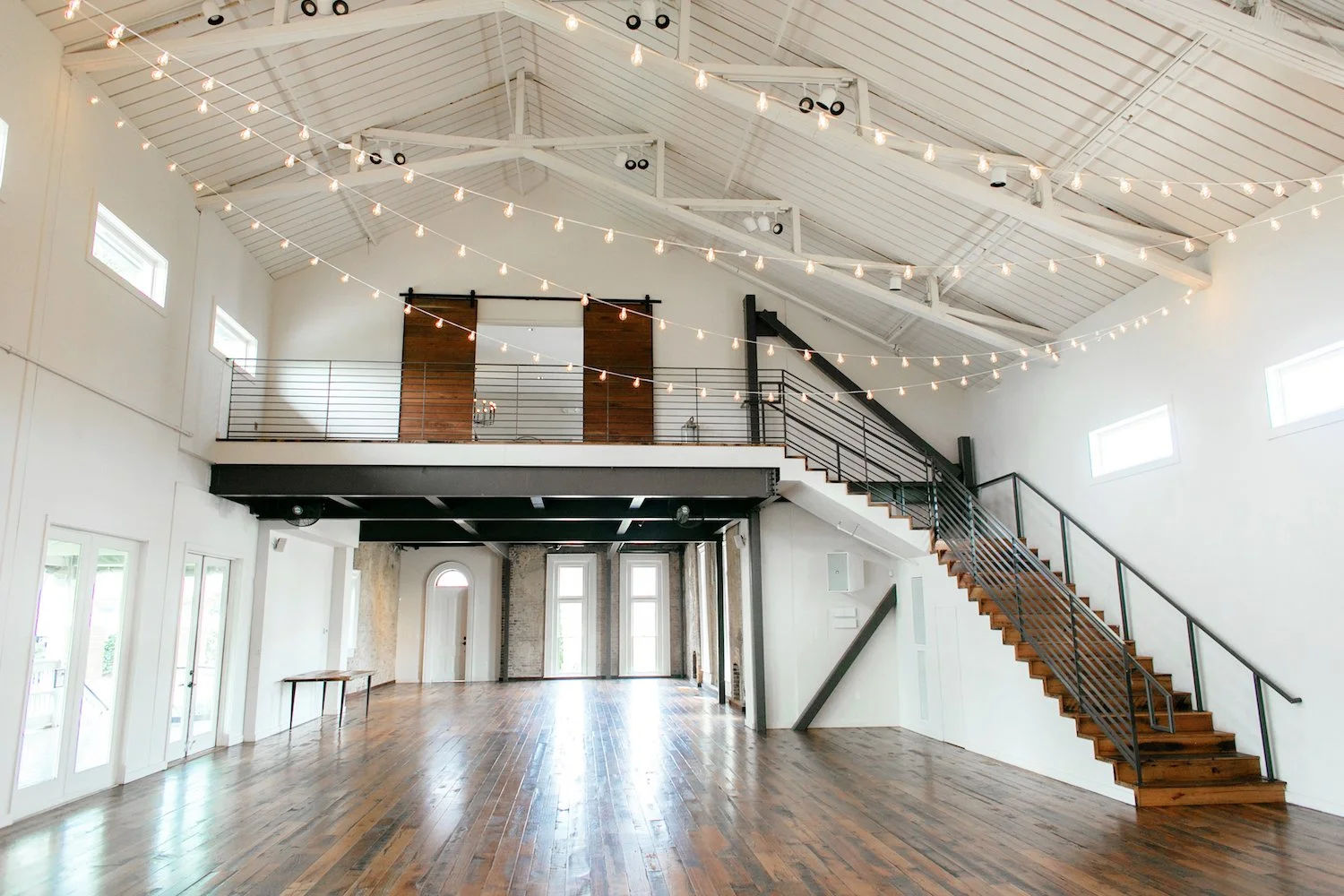 Great Hall and Victorian at the Cordelle in Nashville Interior of a spacious, modern event hall or loft with wooden floors, high ceilings, string lights, windows, a staircase leading to a mezzanine, and minimal furniture.