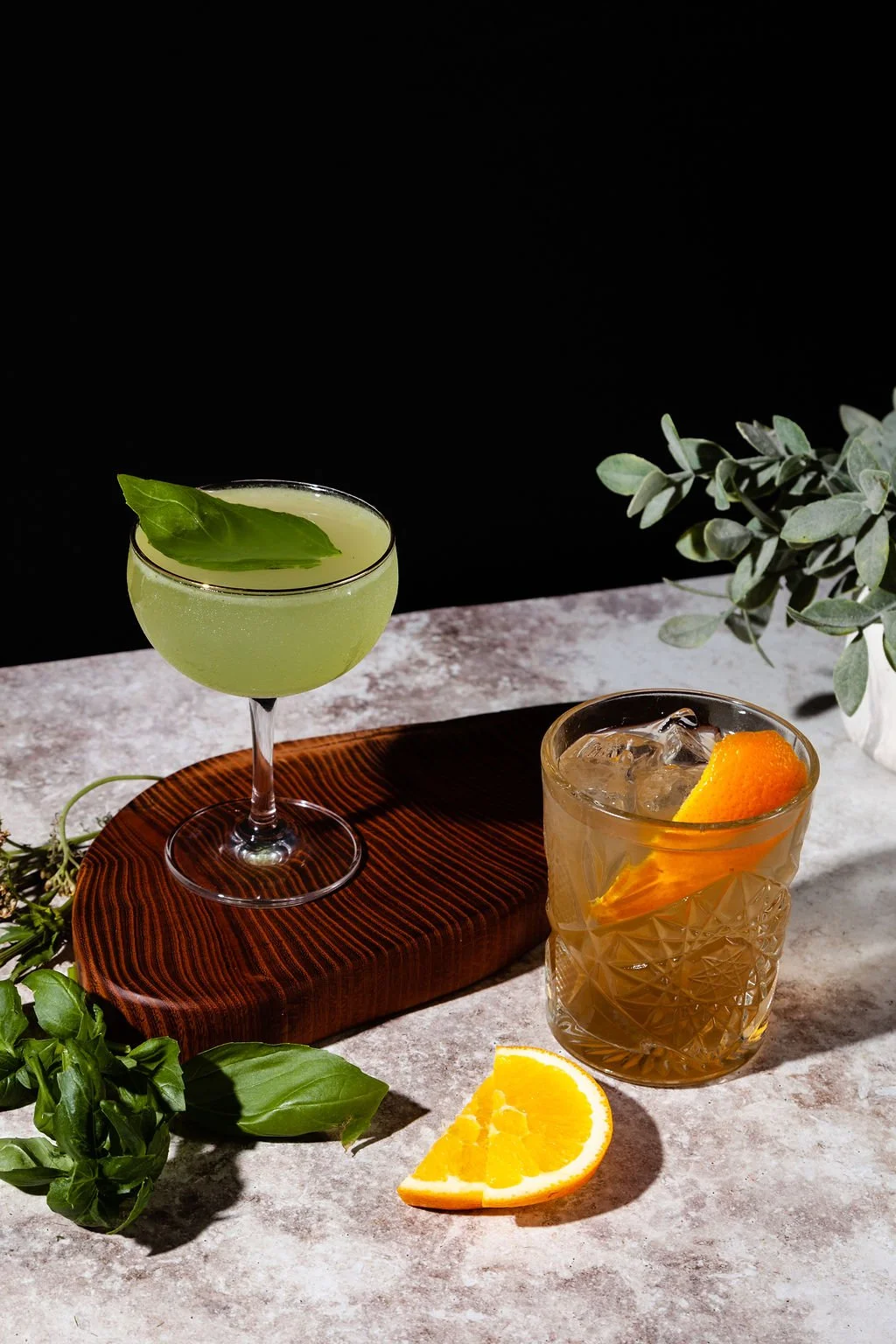 His & Hers Wedding cocktails at the Cordelle on a textured surface with green foliage. One is a light green drink in a coupe glass garnished with a basil leaf. The other is a whiskey-colored drink in a glass with ice and orange slices.