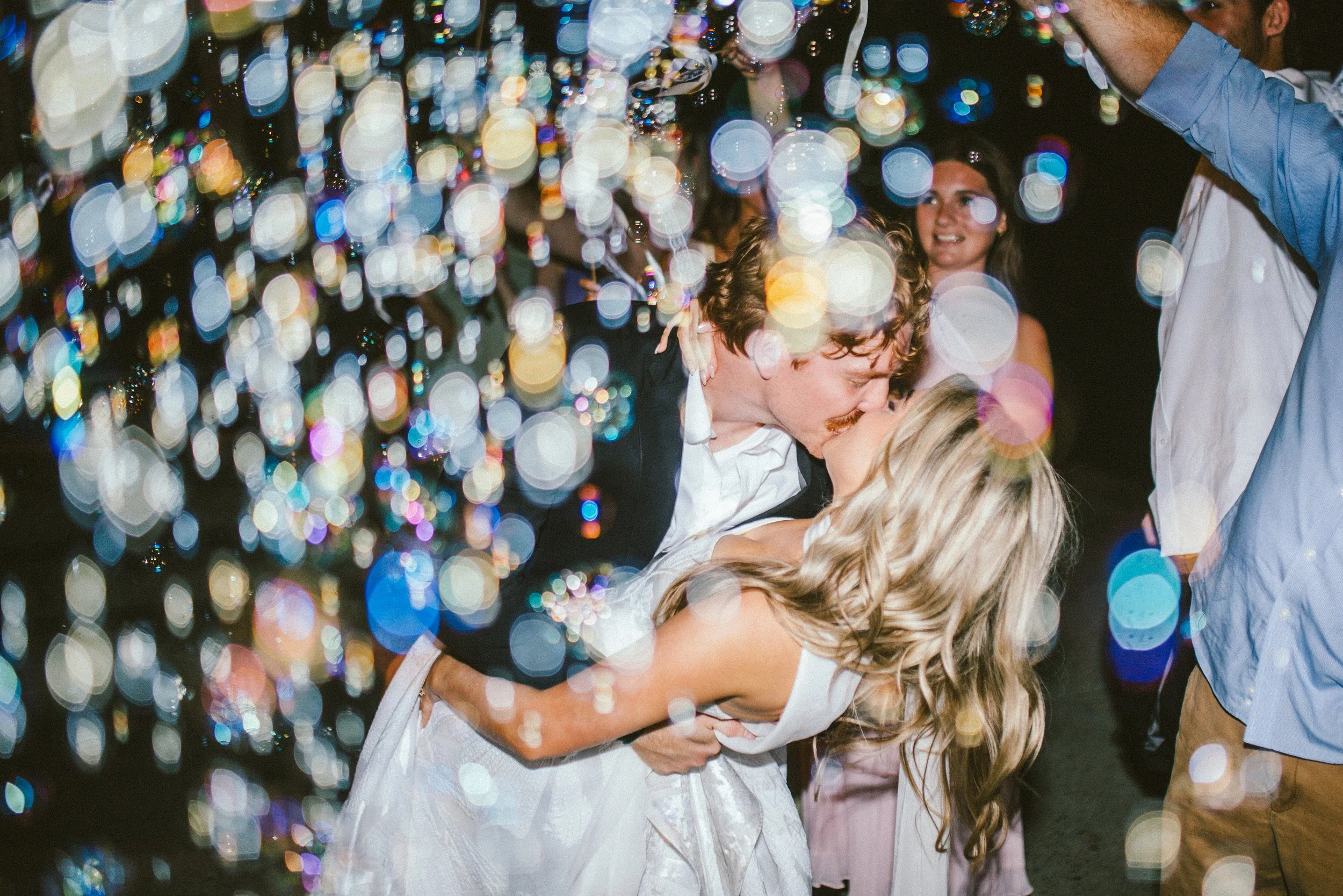 A Cordelle couple send-off of people kissing at a celebration, with colorful lights and blurred background, and a woman smiling in the background.