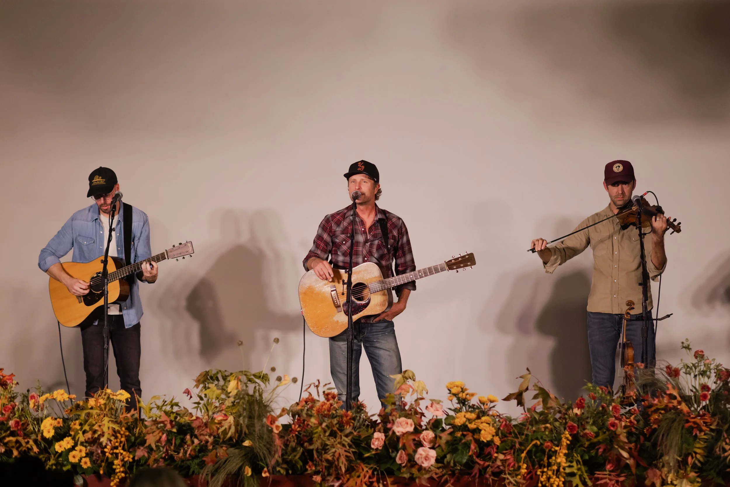 Country music at the Cordelle in Nashville Three men perform on stage with musical instruments, surrounded by colorful flowers, against a plain white backdrop.