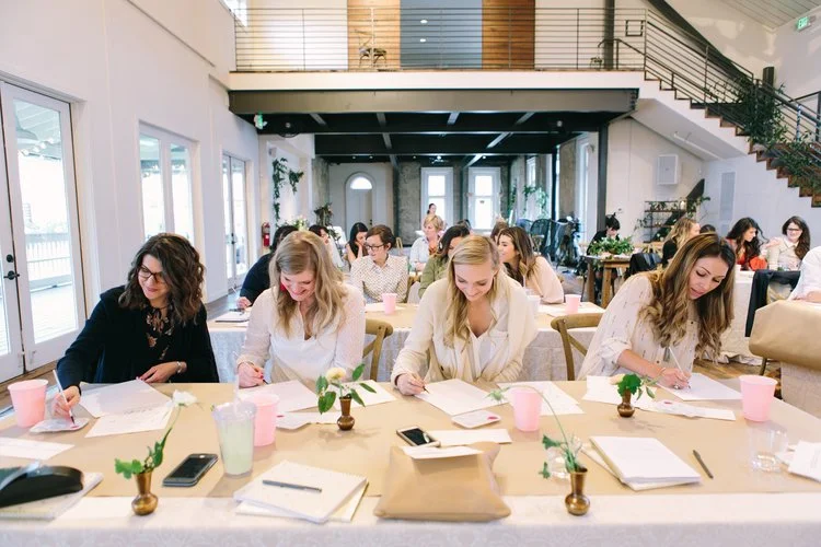Workshop for corporate event at the Cordelle in Nashville Women sitting at a long table in a bright, modern room, working on papers with notebooks, pens, and pink cups, with other people seated at tables in the background.