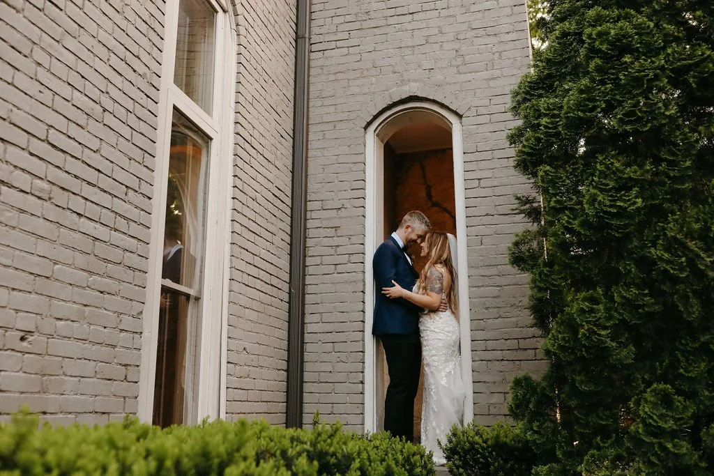 A Cordelle couple sharing an intimate moment in the historic entry way outdoor space next to a brick building, surrounded by greenery.