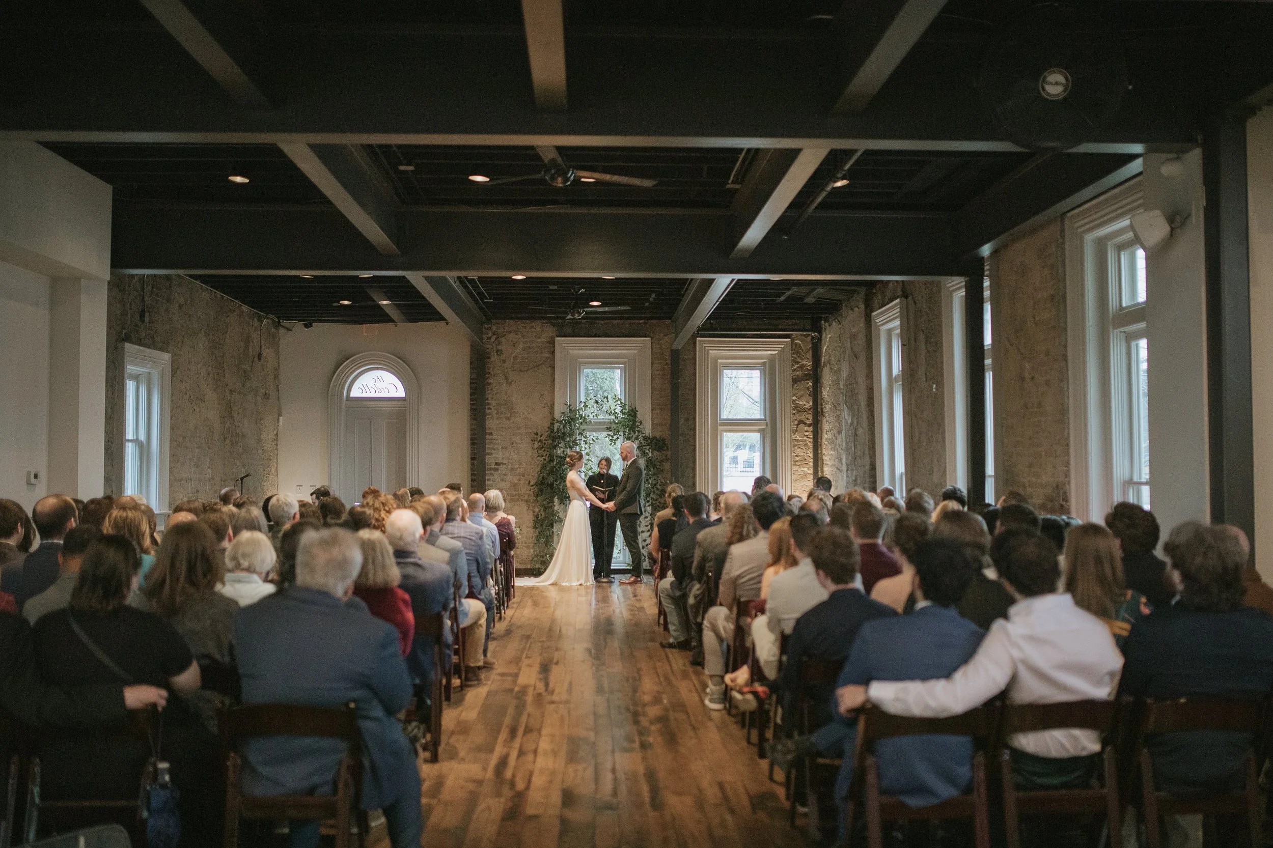 A Cordelle wedding ceremony with an indoor setting featuring exposed brick walls, large windows, and high ceilings. The bride and groom are holding hands and exchanging vows in front of an officiant, with many guests seated and watching.